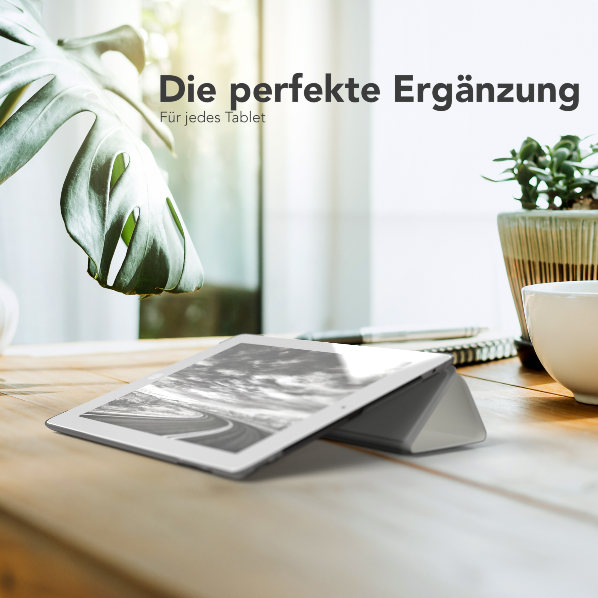 Ein Tablet liegt auf einem Holztisch und ist aufgestellt. Ein großes grünes Blatt und eine kleine Pflanze im Topf sind im Hintergrund.