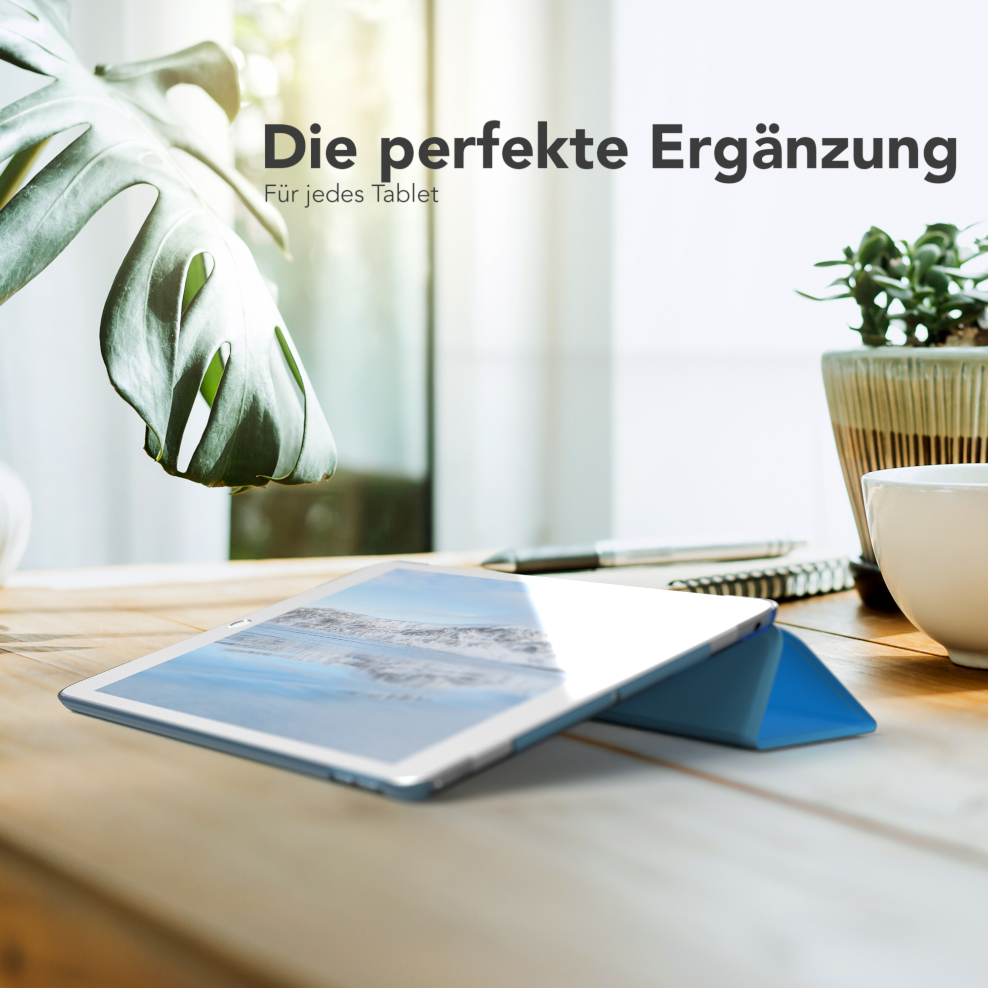 Tablet mit blauem Cover, auf einem Holztisch, neben einer Pflanze und einer weißen Tasse. Fenster im Hintergrund.
