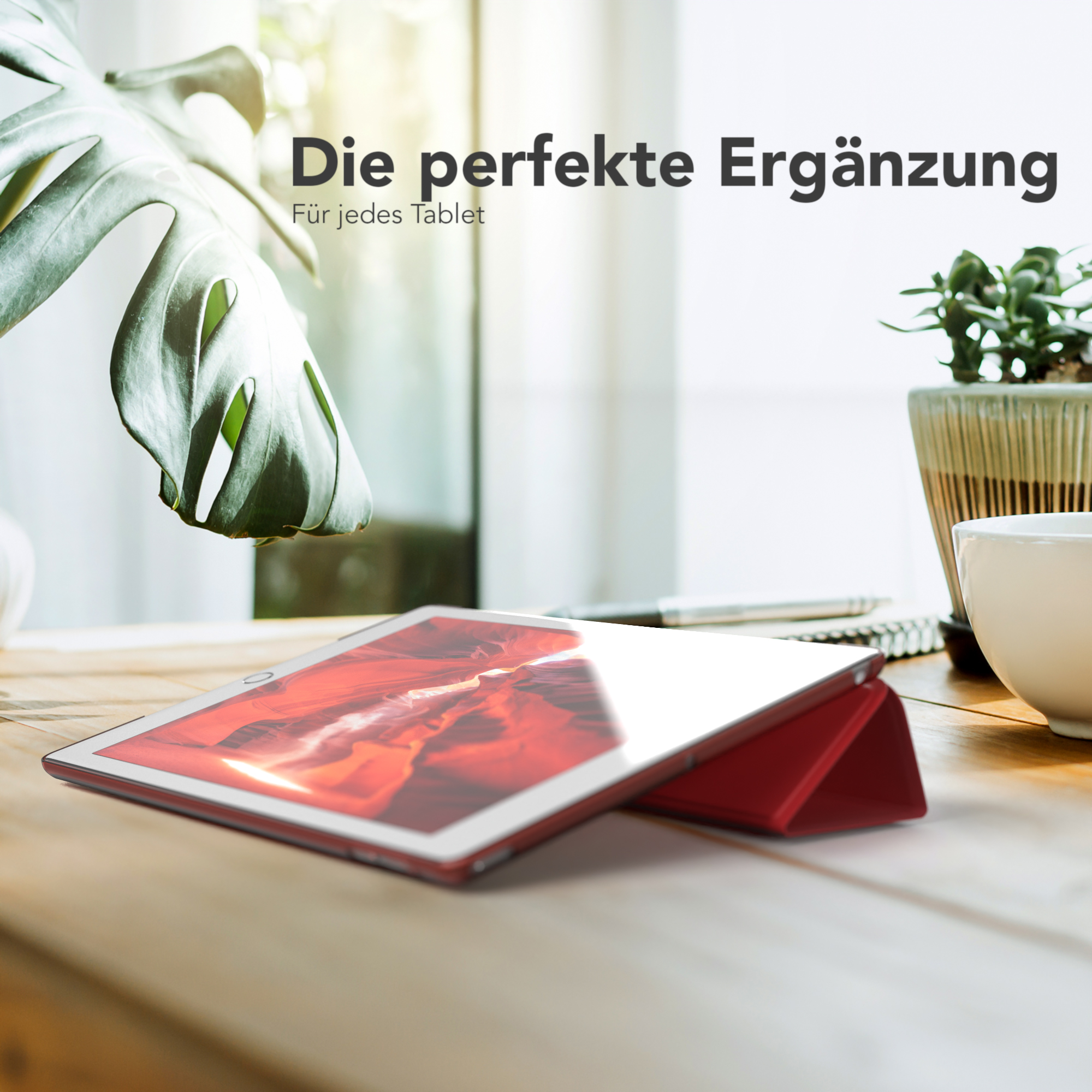 Tablet in roter Hülle auf Holztisch. Grüne Pflanze und Becher im Hintergrund. Helle Beleuchtung.