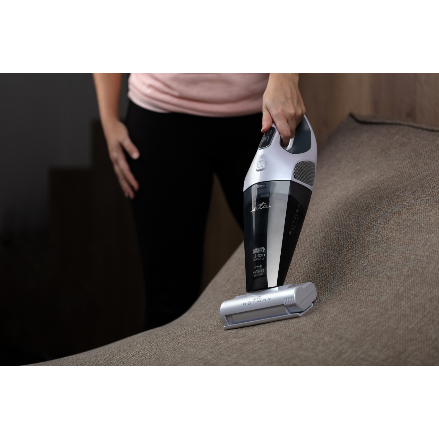 Eine Person reinigt ein Sofa mit einem weiß-schwarzen Handstaubsauger. Braunes Sofa im Hintergrund.