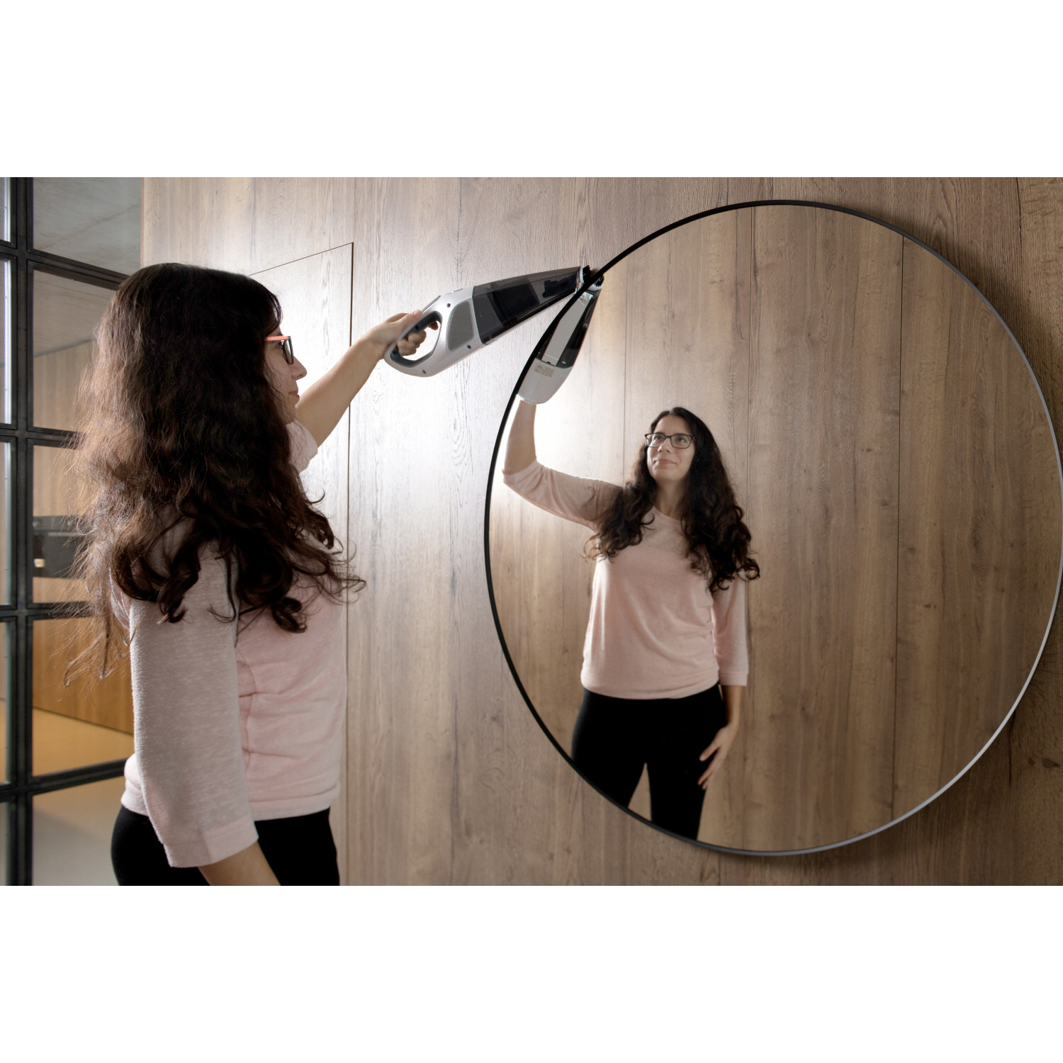 Mujer limpiando un espejo, reflejándose en él, con una aspiradora.