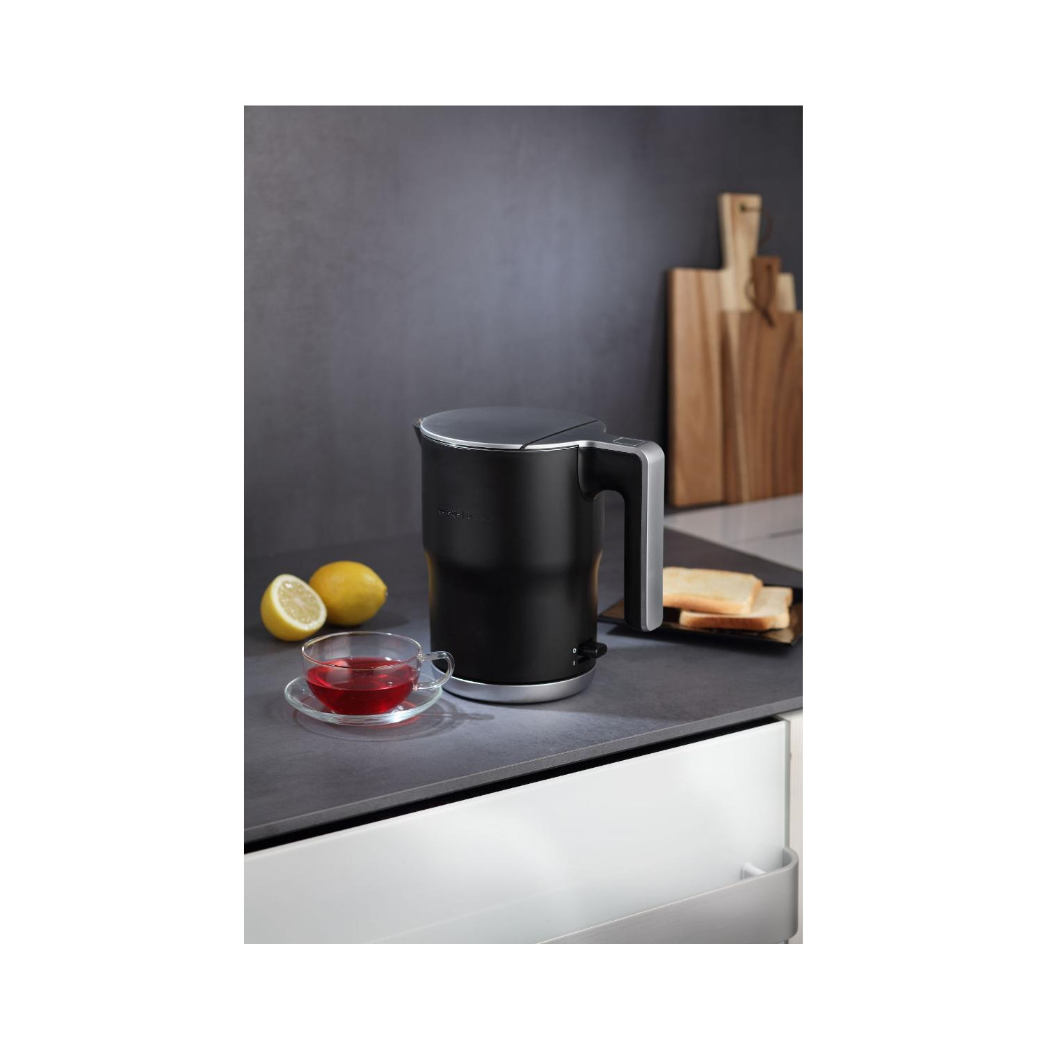 Ein schwarzer Wasserkocher, eine Teetasse mit Tee, eine Zitronenscheibe und Toast auf einer Theke.