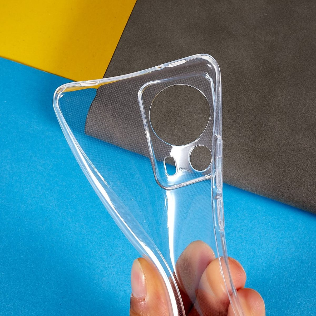 Klar Handyhülle, gehalten von Fingern, vor blauem, gelbem und grauem Hintergrund.