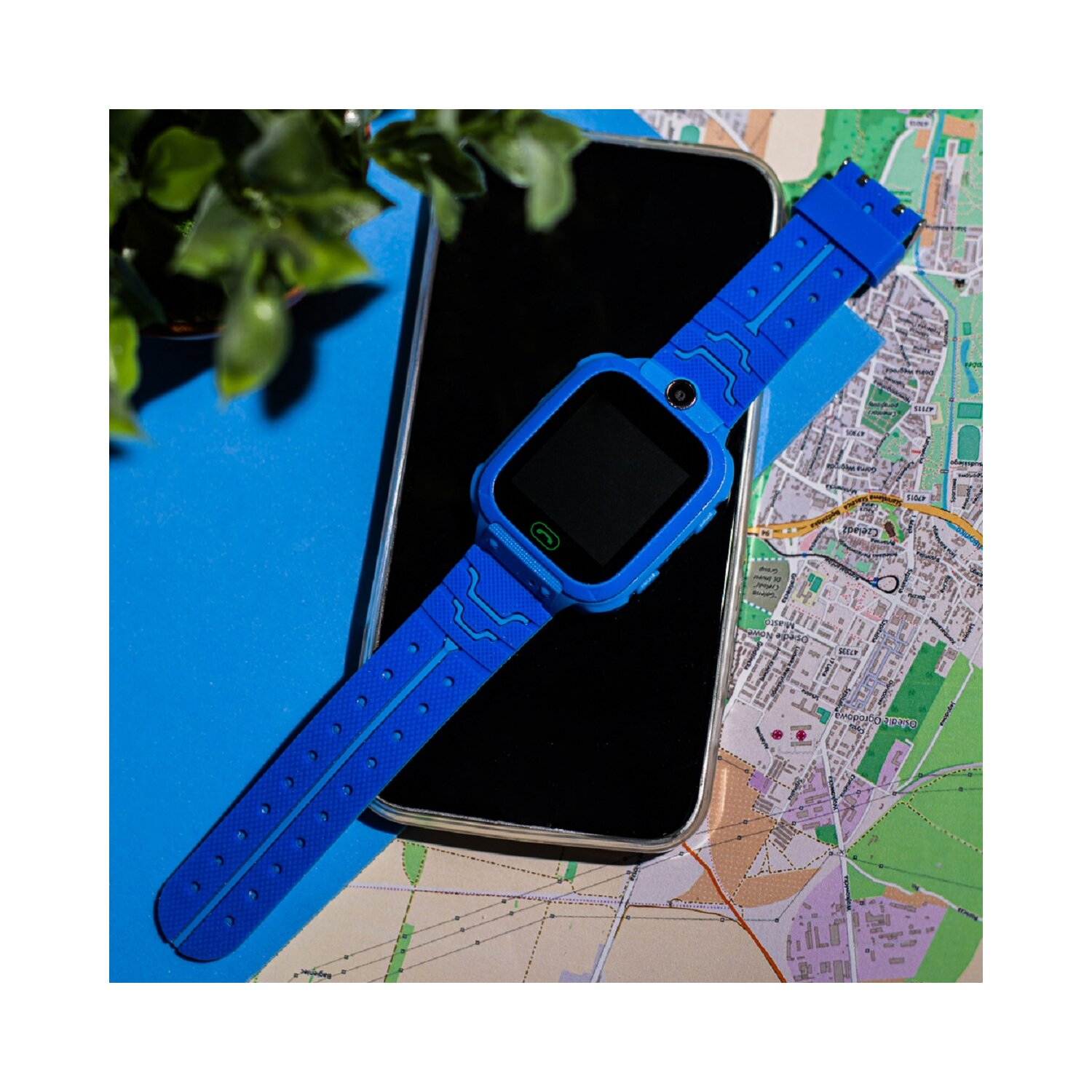 Eine blaue Smartwatch auf einem Telefon und einer Karte. Grüne Pflanze links.