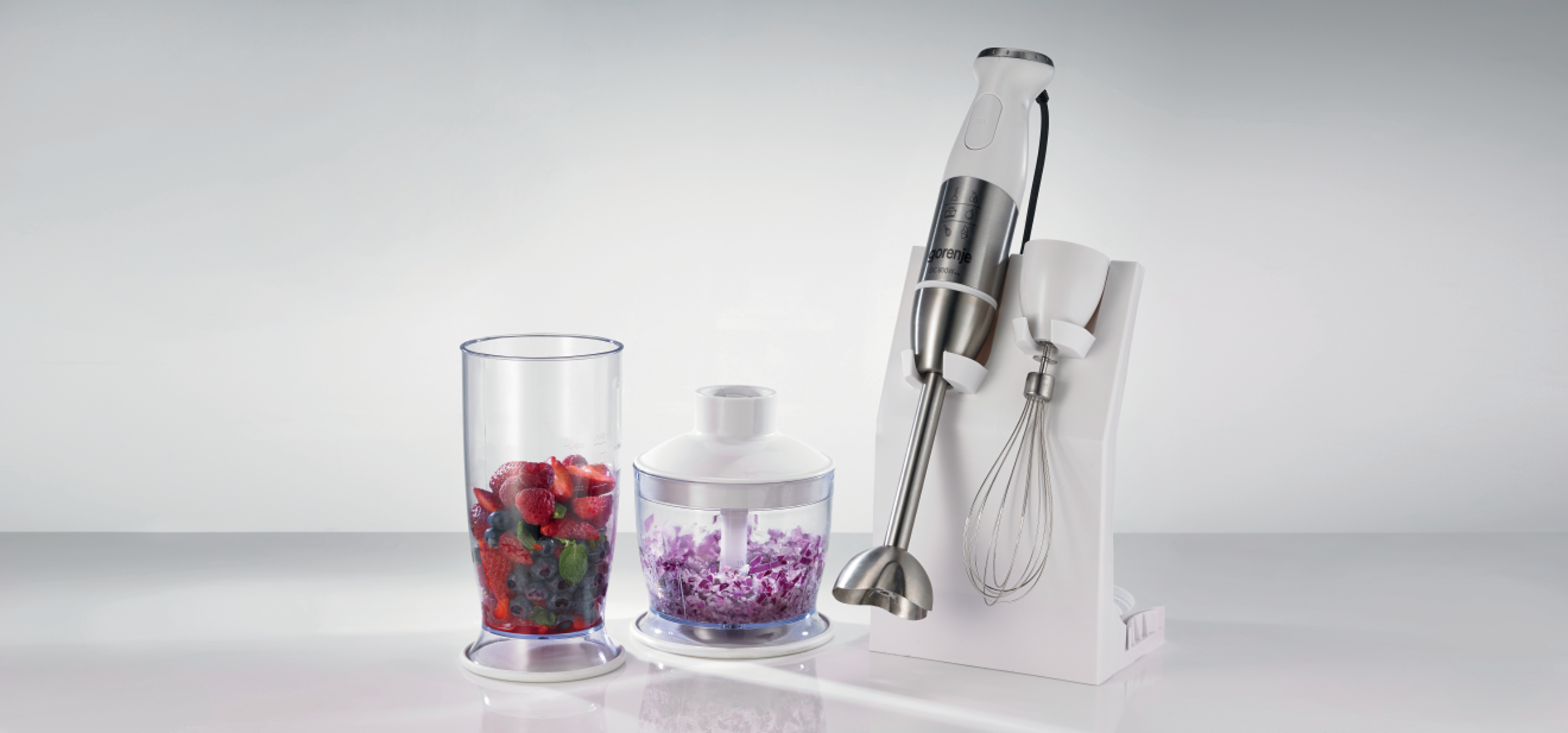 Blender-Set: Mixer, Behälter mit Beeren, Behälter mit gehacktem Gemüse, Schneebesen in weißem Ständer.