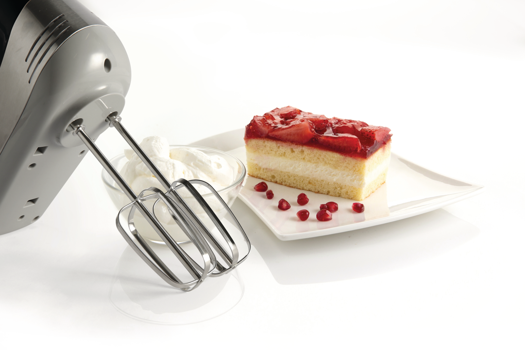 Handrührgerät mit Schneebesen, Schüssel mit Schlagsahne, Kuchenstück mit rotem Gelee und Granatapfelkernen.