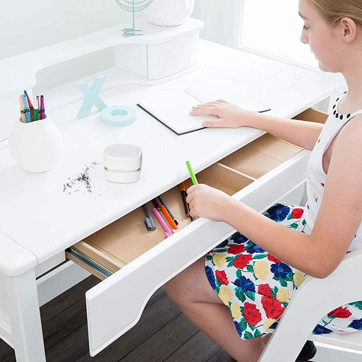 Dziewczyna przy białym biurku, otwierająca szufladę. Szuflada zawiera długopisy i małe białe pudełko.
