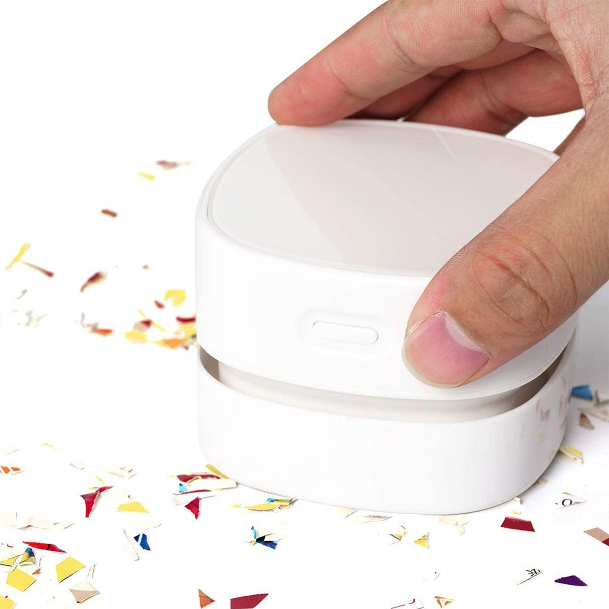 Biały mini odkurzacz z papierowym konfetti na białej powierzchni, trzymany przez dłoń.