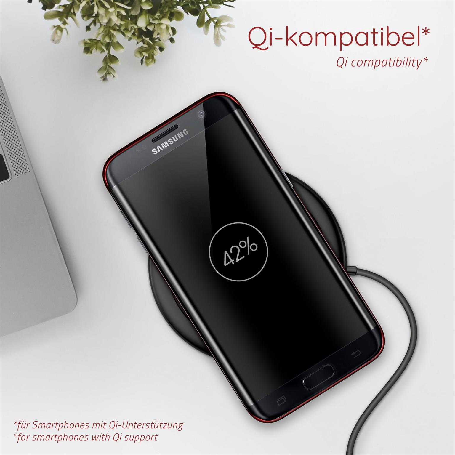 Schwarzes Smartphone auf einem kabellosen Ladegerät. Der Bildschirm zeigt 42%. Im Hintergrund ein Laptop und eine Pflanze.