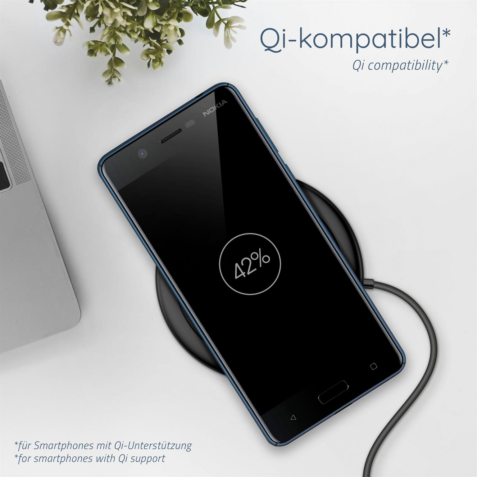 Schwarzes Telefon, das drahtlos auf einem Ladepad geladen wird, mit Pflanze und Laptop im Hintergrund.
