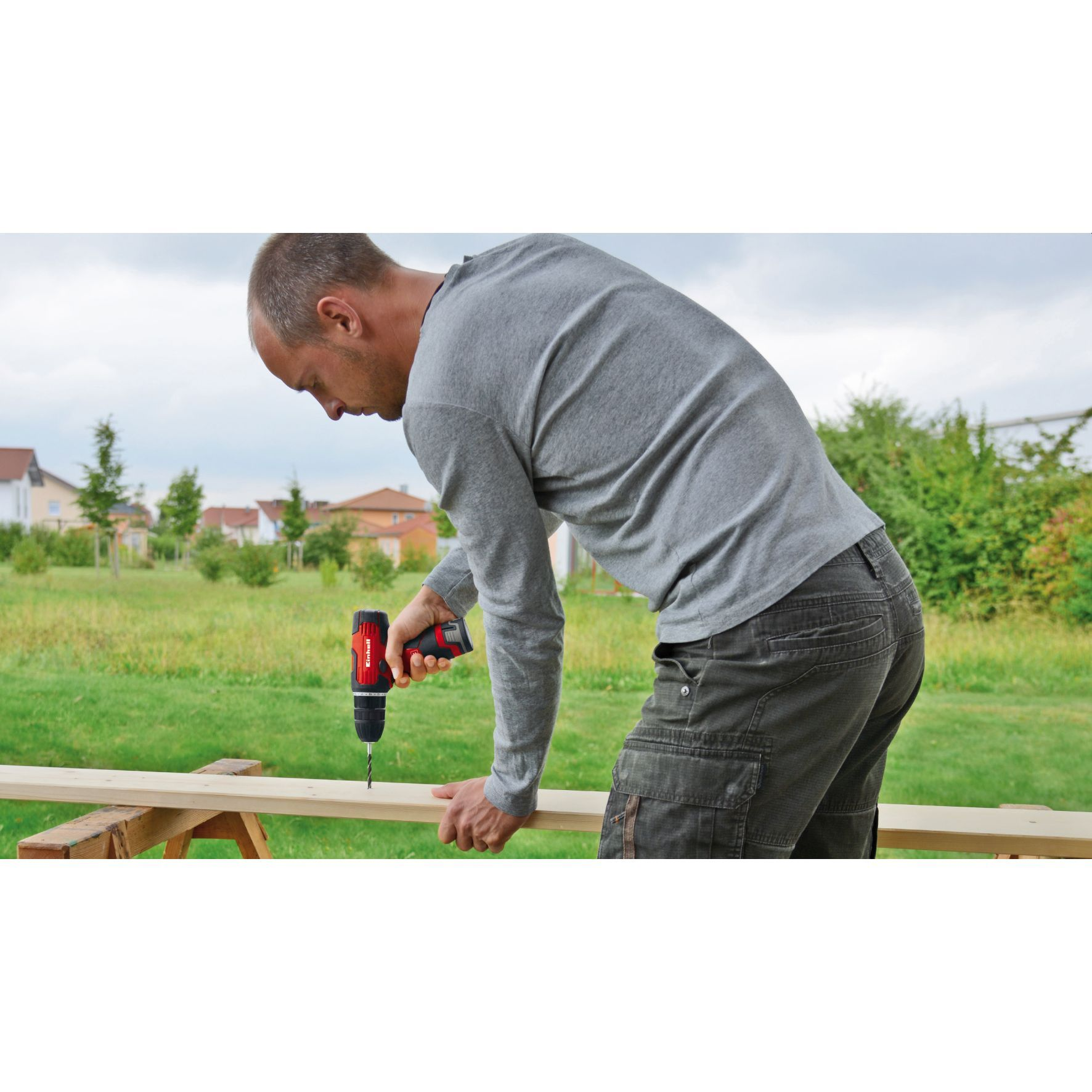 Mężczyzna wierci w drewnie. Ma na sobie szarą koszulę i spodnie cargo, pracuje na zewnątrz w pobliżu domów.