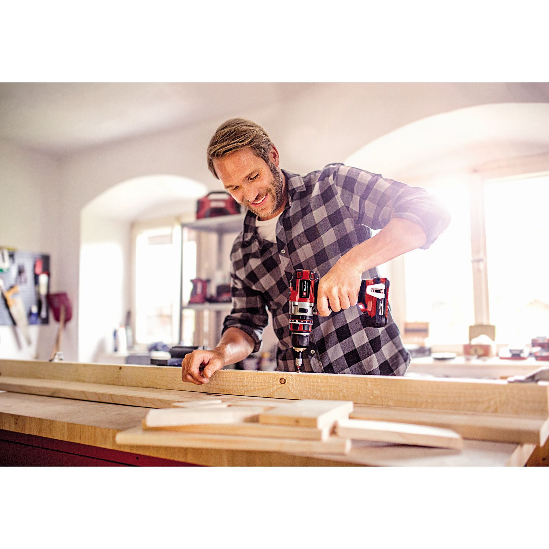 Mężczyzna wierci w drewnie. Ma na sobie koszulę w kratę. Warsztat, narzędzia i okno w tle.