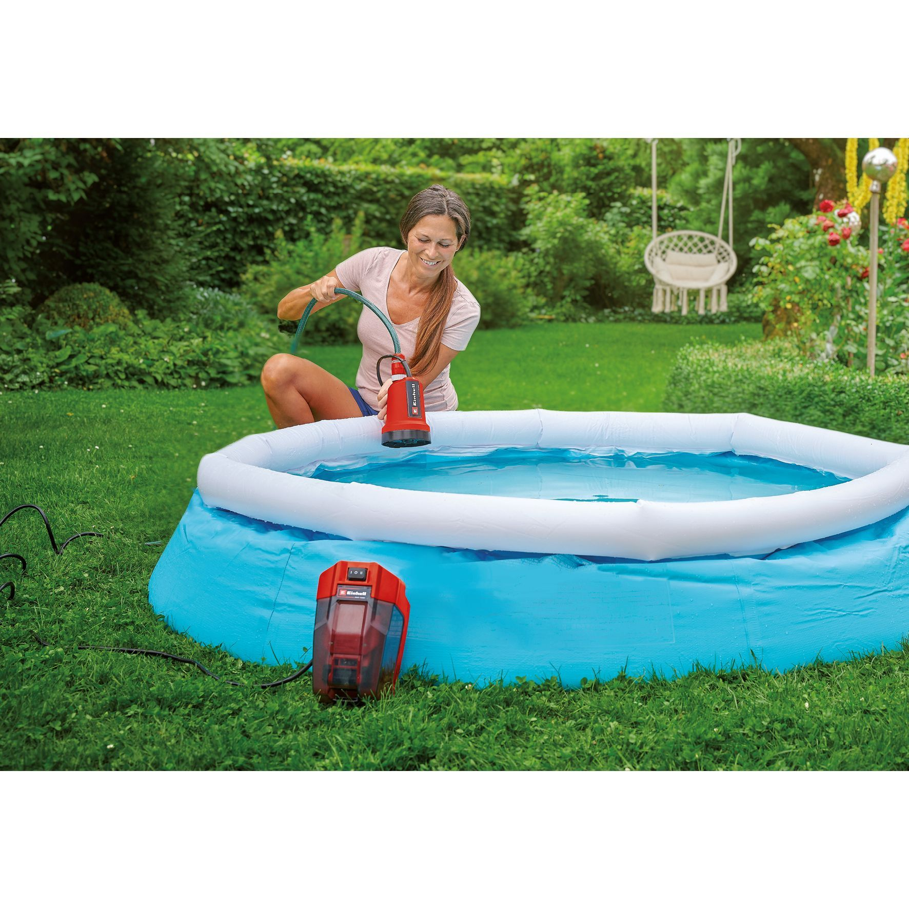 Kobieta na podwórku napełnia niebieski basen pompą. Pompa jest czerwono-czarna. Otacza ją zielona trawa.