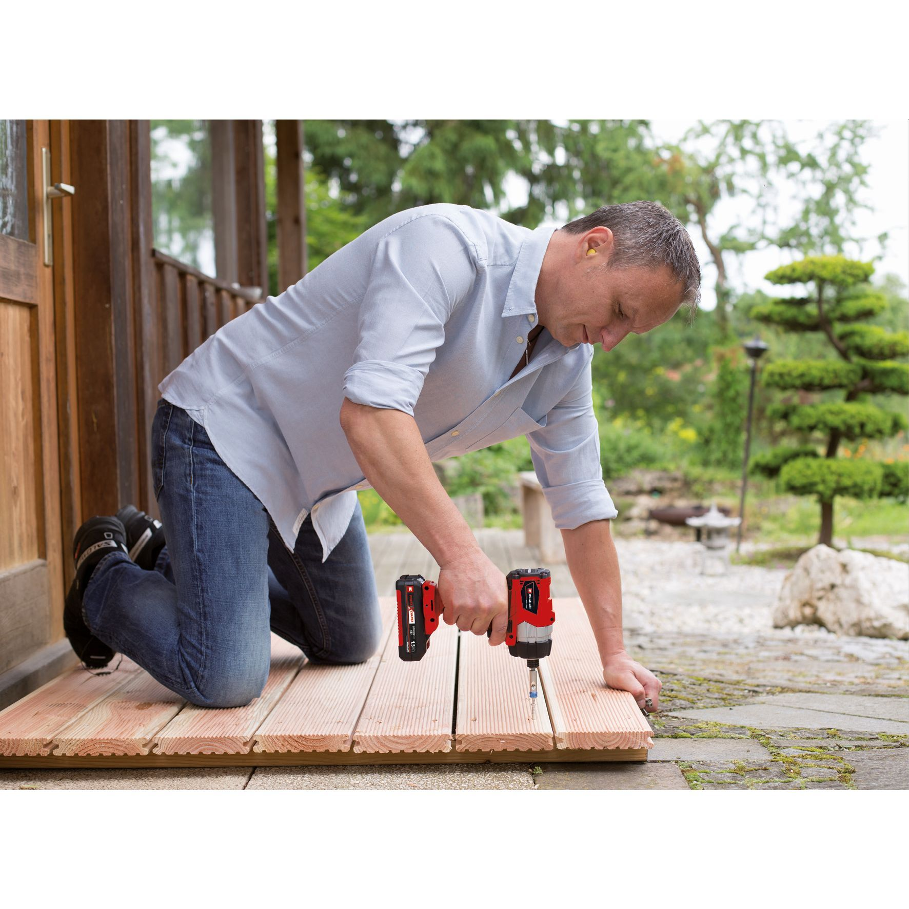 Mężczyzna w dżinsach i koszuli klęczy, używając czerwonej wiertarki na drewnianych deskach.