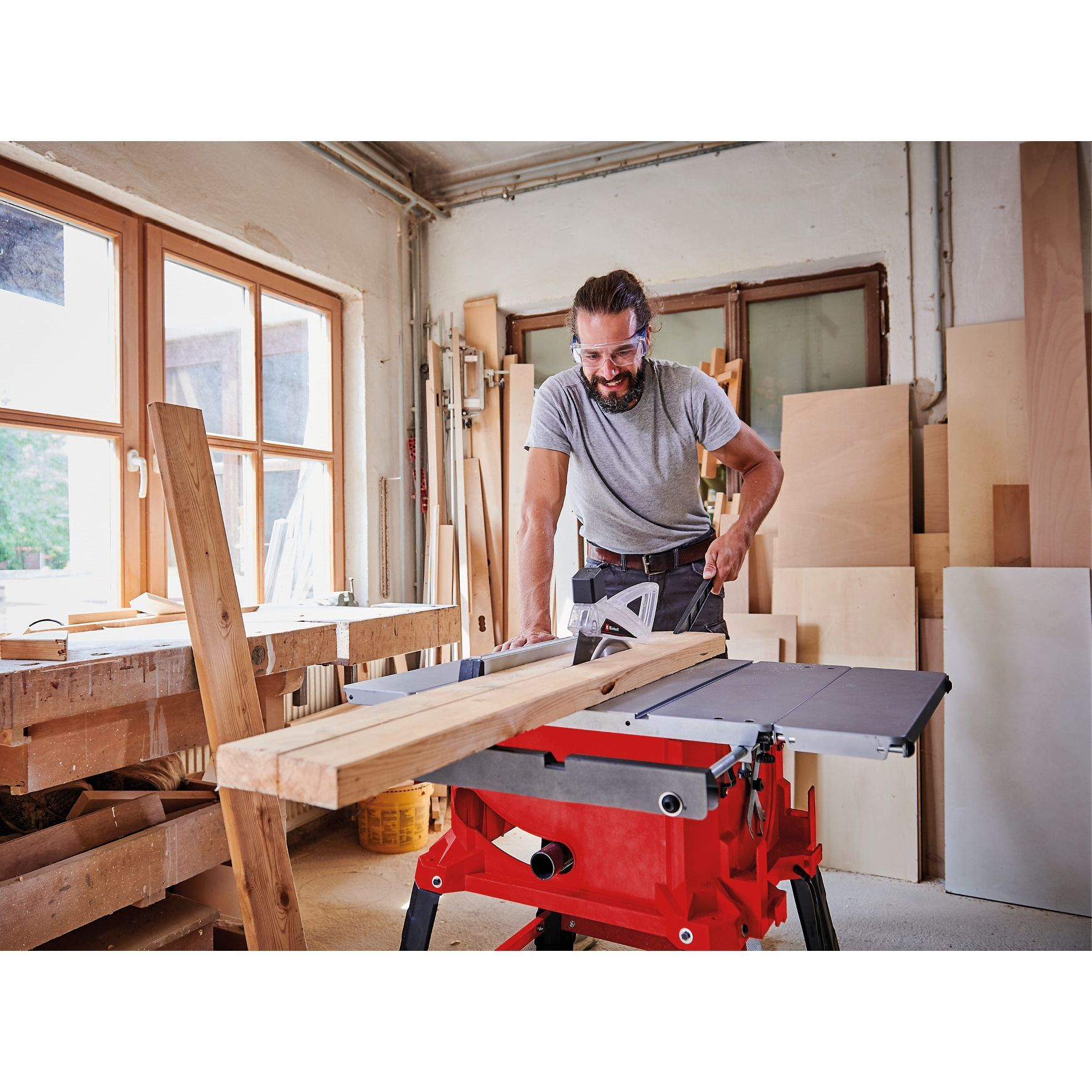 Mężczyzna w okularach ochronnych tnie drewno piłą stołową w warsztacie.