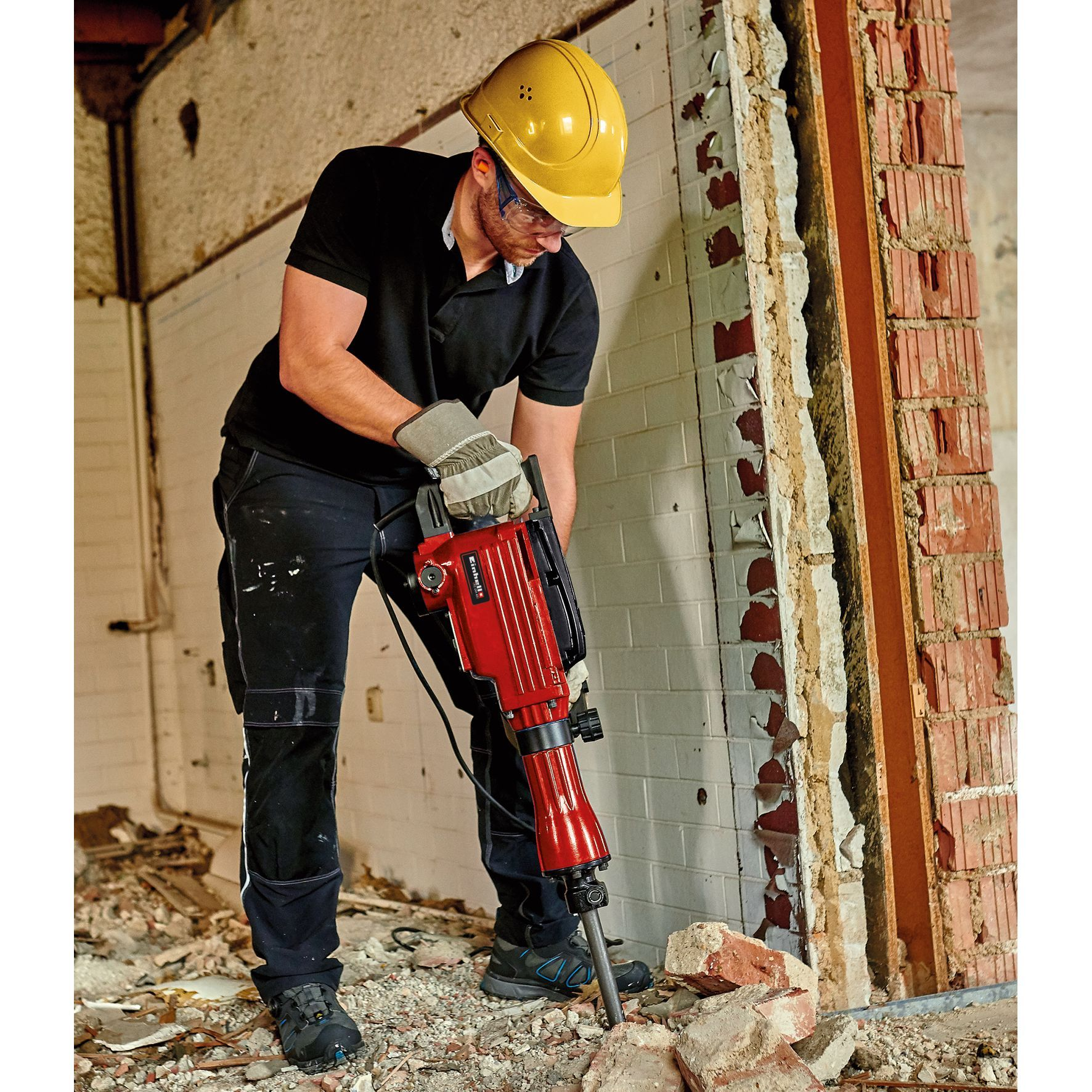 Mężczyzna w żółtym kasku i okularach ochronnych używa czerwonego młota wyburzeniowego do rozbiórki ściany.