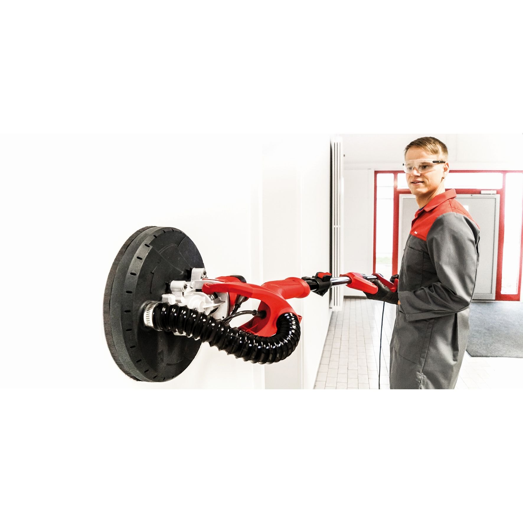 Homme ponçant un mur avec une ponceuse rouge et noire, portant des lunettes de sécurité.