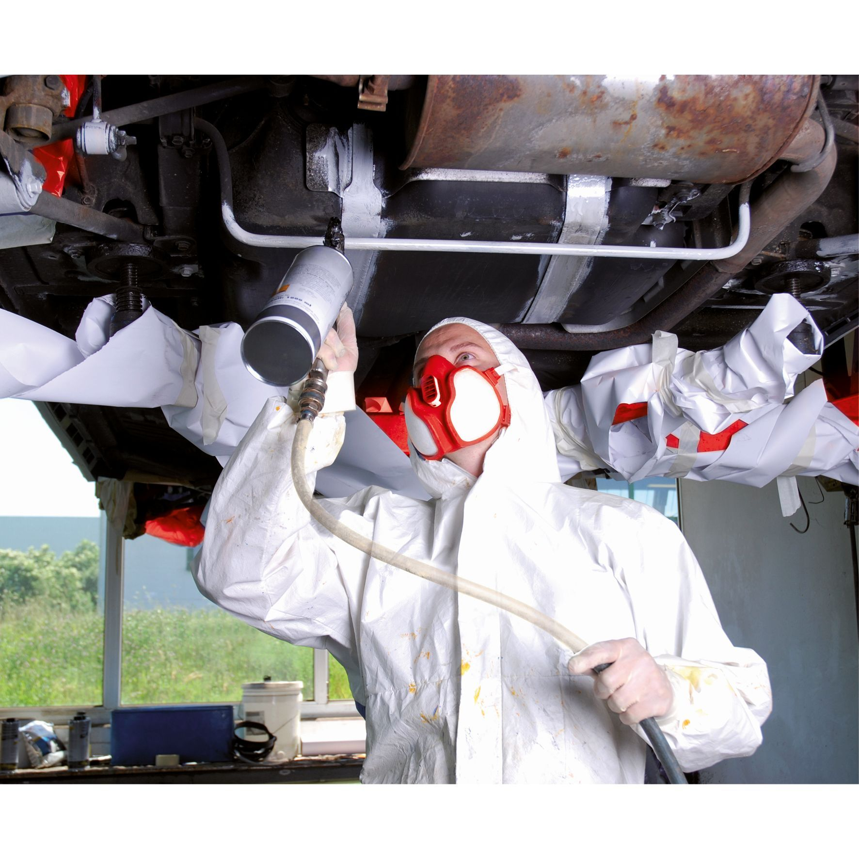 Eine Person in einem weißen Schutzanzug und Maske sprüht Farbe auf die Unterseite eines Autos.