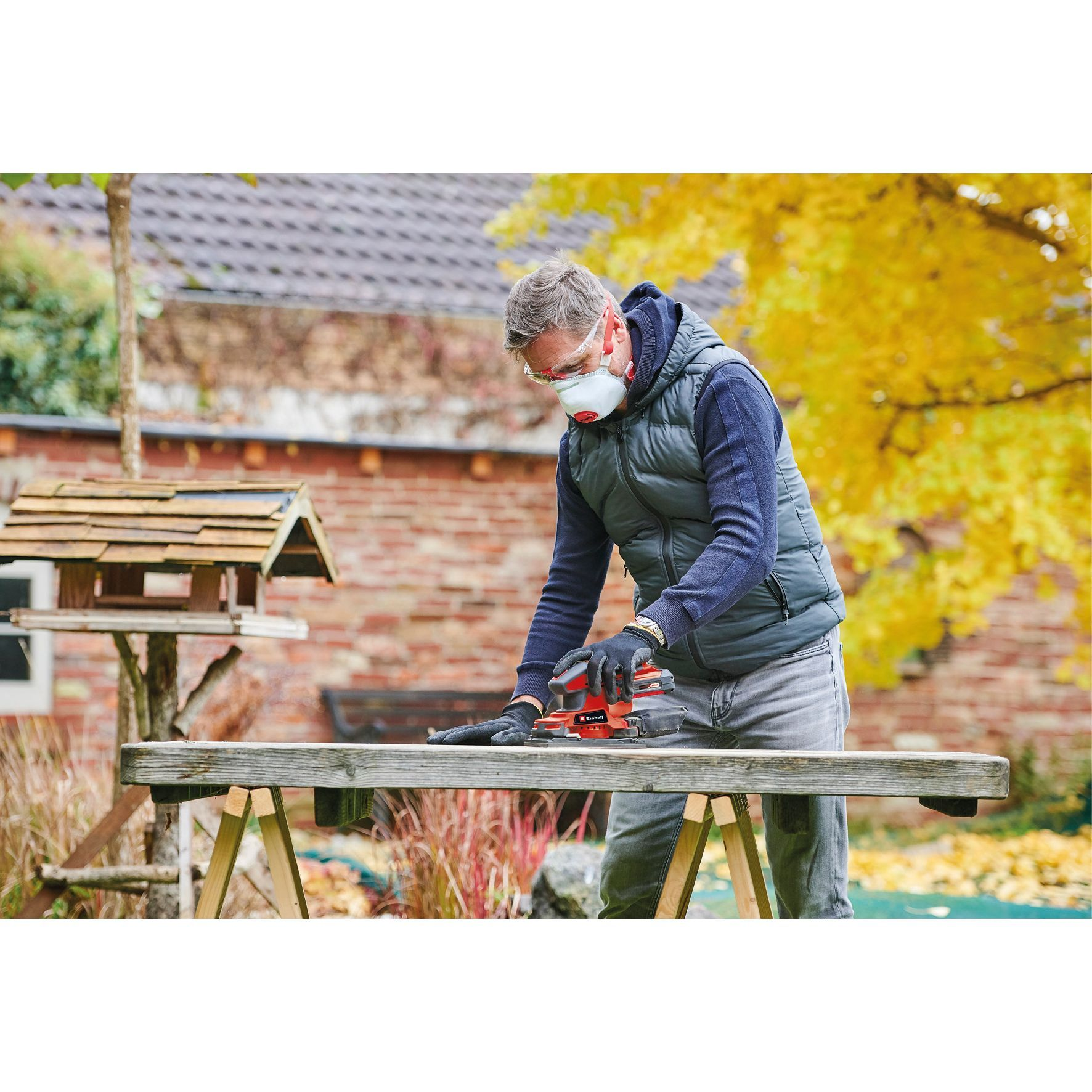 Mężczyzna szlifujący drewno na zewnątrz czerwoną szlifierką, w masce i rękawicach.