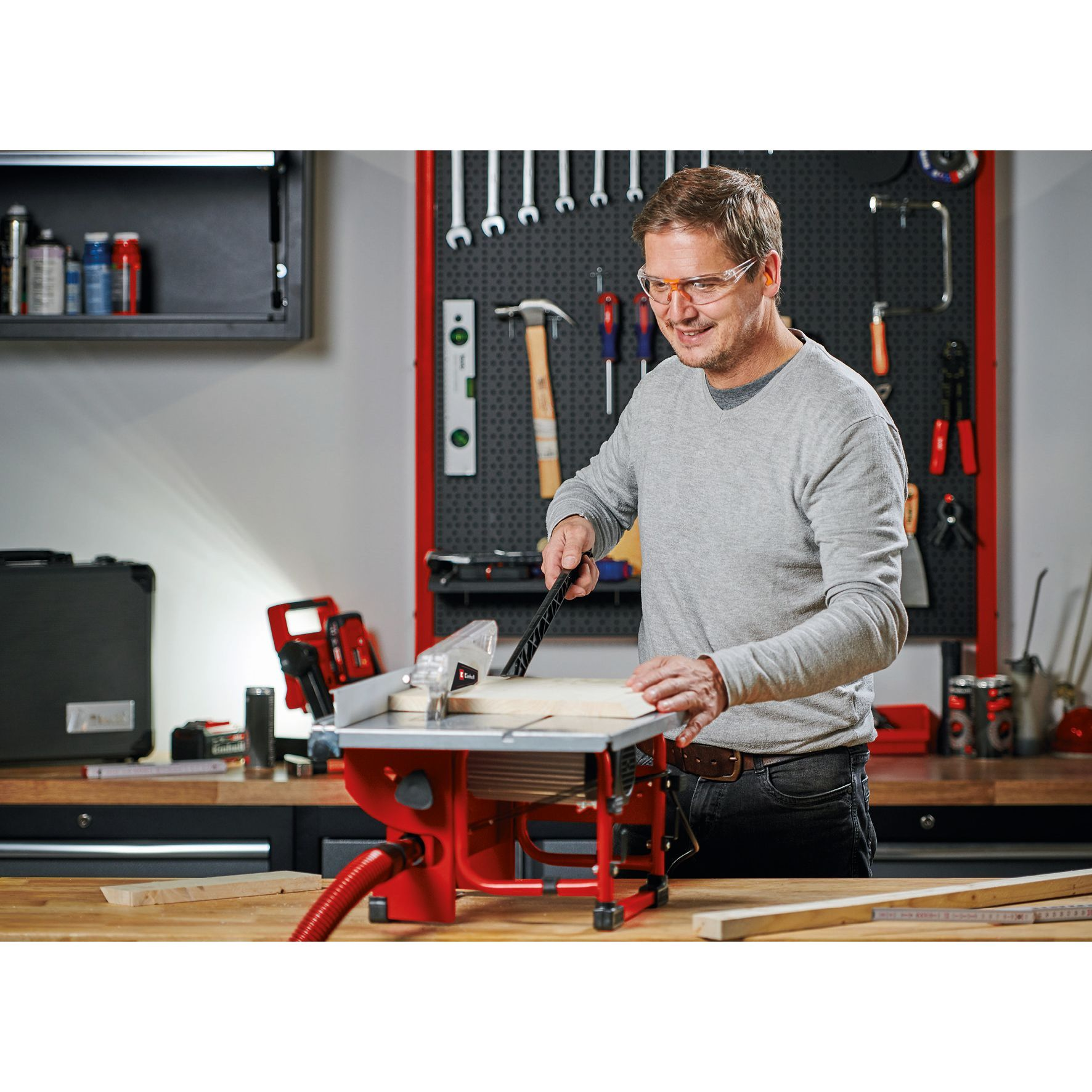 Mann schneidet Holz mit einer roten Tischkreissäge. Werkstatt-Hintergrund mit Werkzeugen und Lochwand.