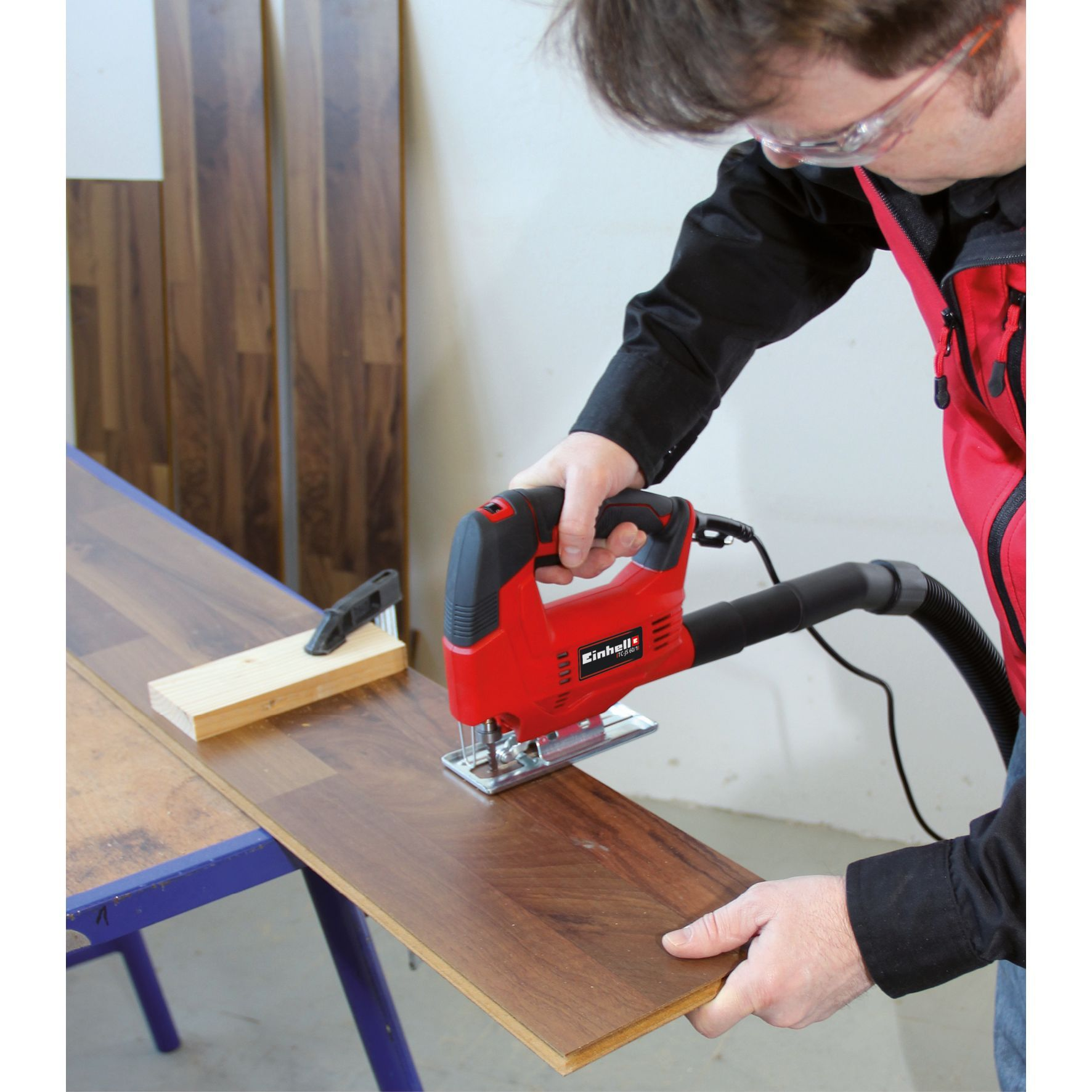 Mężczyzna tnący drewno czerwoną wyrzynarką, ma na sobie okulary ochronne.