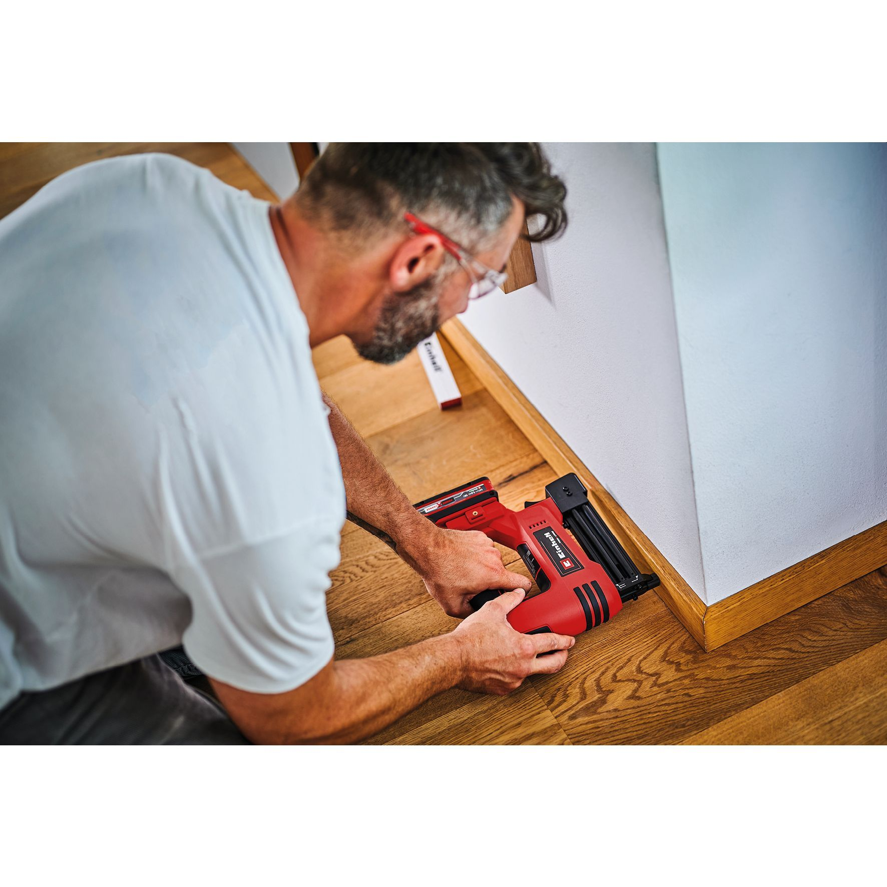 Man die een rode spijkerpistool gebruikt op een houten frame bij een muur.