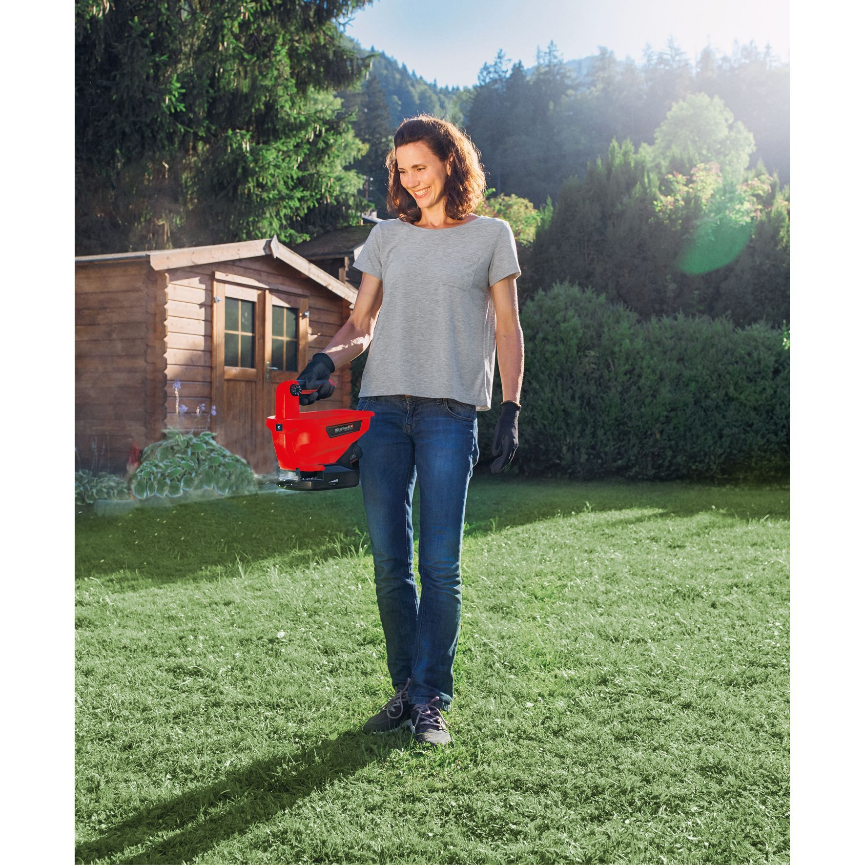Kobieta w dżinsach i szarej koszulce trzymająca czerwone elektronarzędzie, stojąca na trawie w ogrodzie z szopą.
