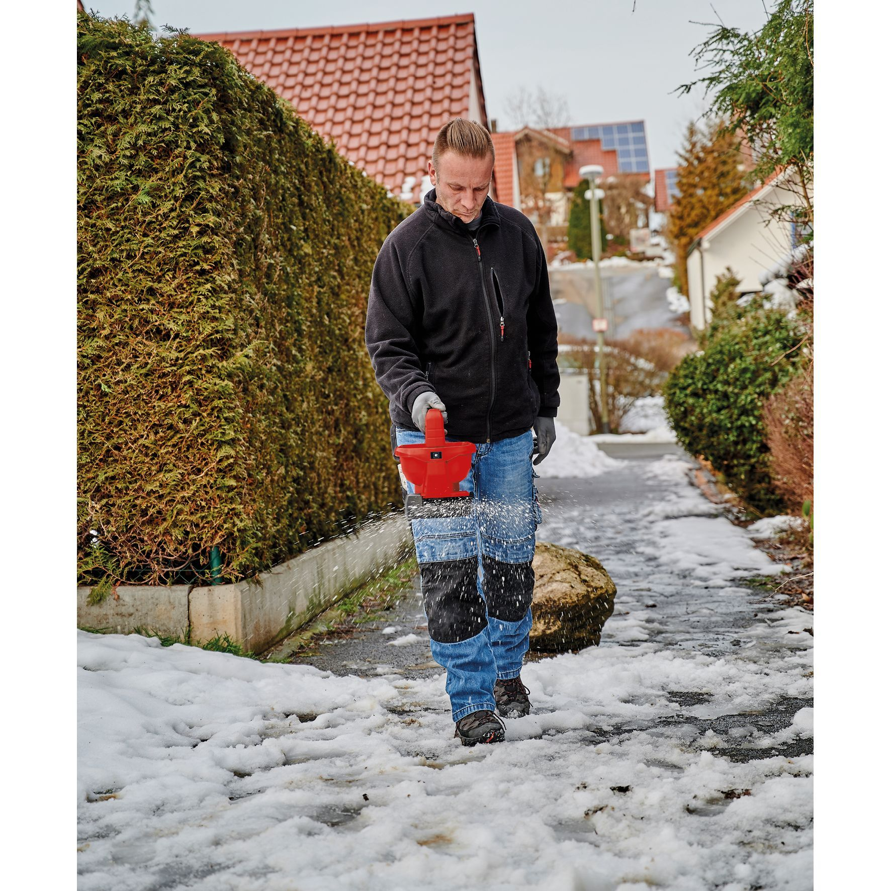 Mężczyzna idący po zaśnieżonej ścieżce, rozsypujący sól. Ubrany w dżinsy, czarną kurtkę i rękawice.