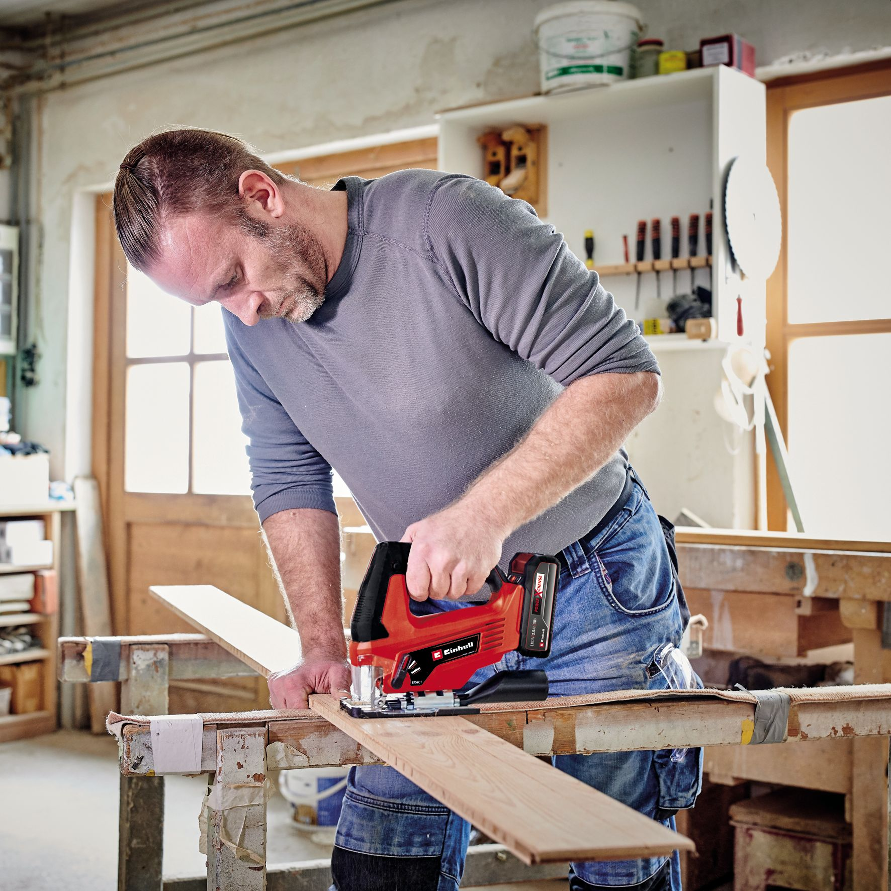 Ein Mann benutzt eine rote Stichsäge, um Holz in einer Werkstatt zu schneiden, trägt ein graues Hemd und Jeans.