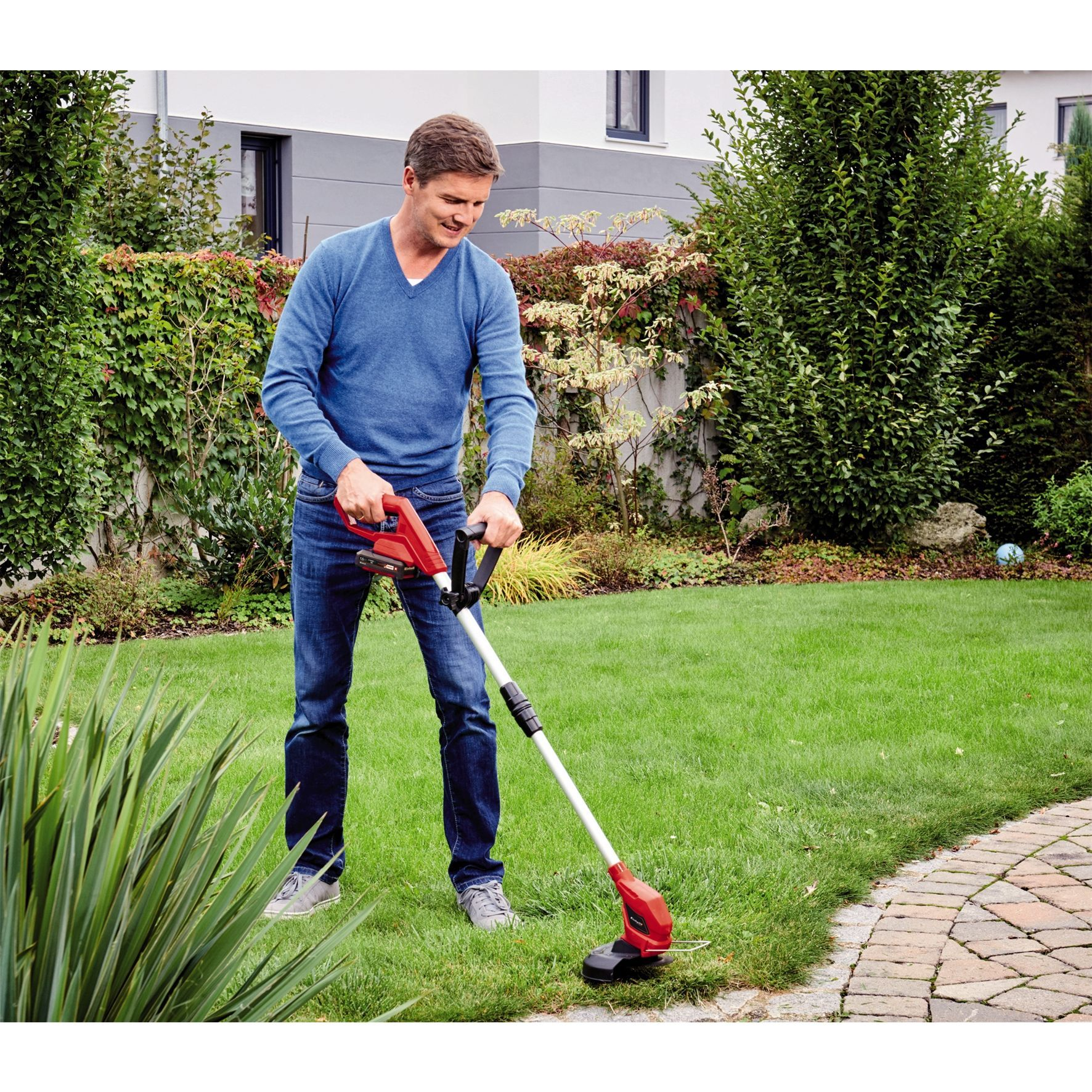 Ein Mann benutzt einen rot-schwarzen Trimmer auf einem Rasen. Grünes Gras, Büsche und ein Weg sind sichtbar.