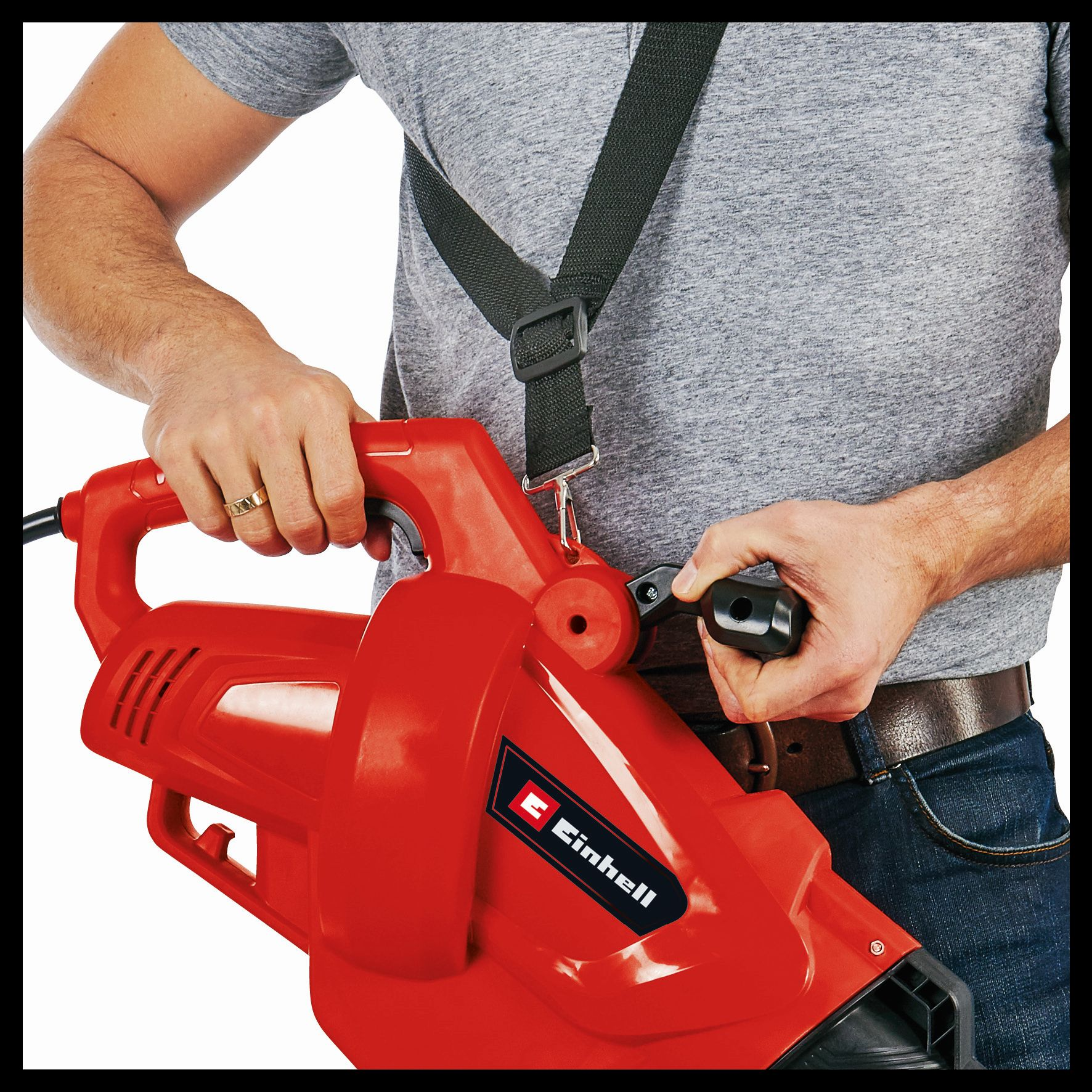 Mężczyzna trzymający czerwoną dmuchawę do liści. Szara koszula i dżinsy. Czarny pasek.