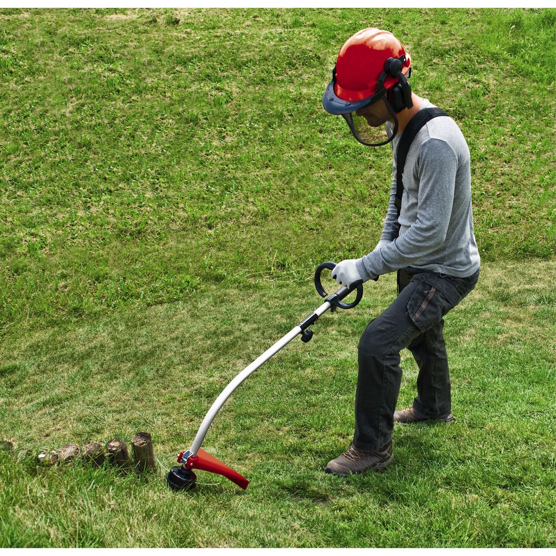 Mann mit Schutzkleidung benutzt einen Trimmer auf einer Wiese. Hintergrund zeigt einen Baumstumpf.