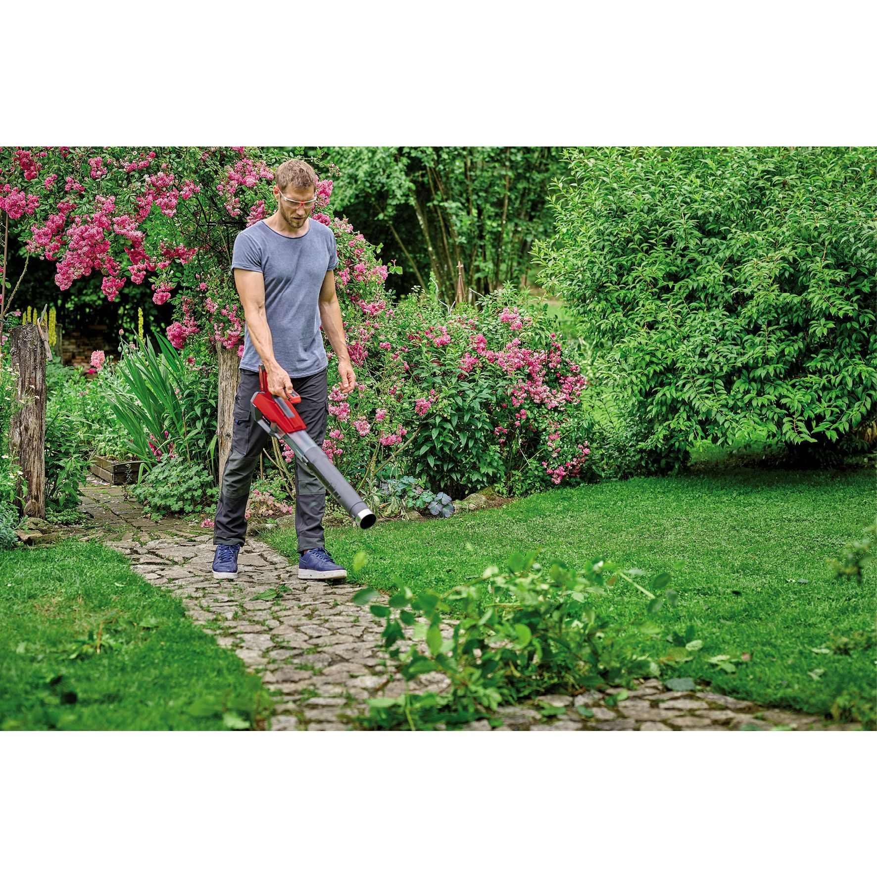 Mężczyzna używający dmuchawy do liści do usuwania liści w ogrodzie z kwiatami i trawą.