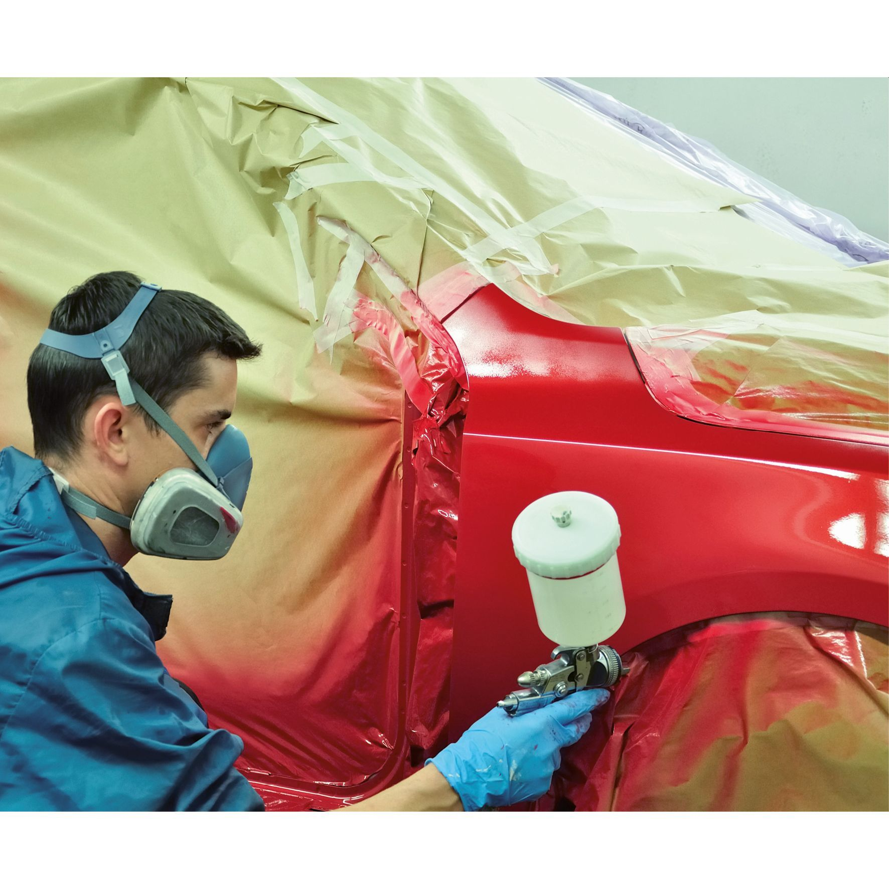 Ein Mann mit Atemschutzmaske sprüht rote Farbe auf ein rotes Auto in einer Werkstatt.