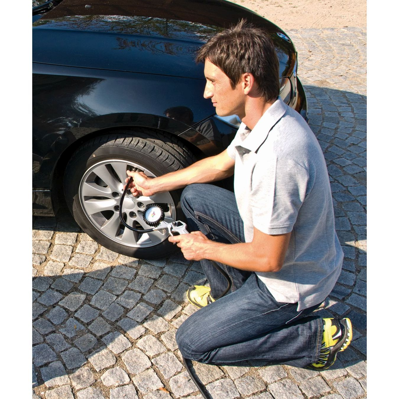 Mann kniet, pumpt einen Autoreifen mit einem Manometer auf. Er trägt Jeans und ein graues Hemd.