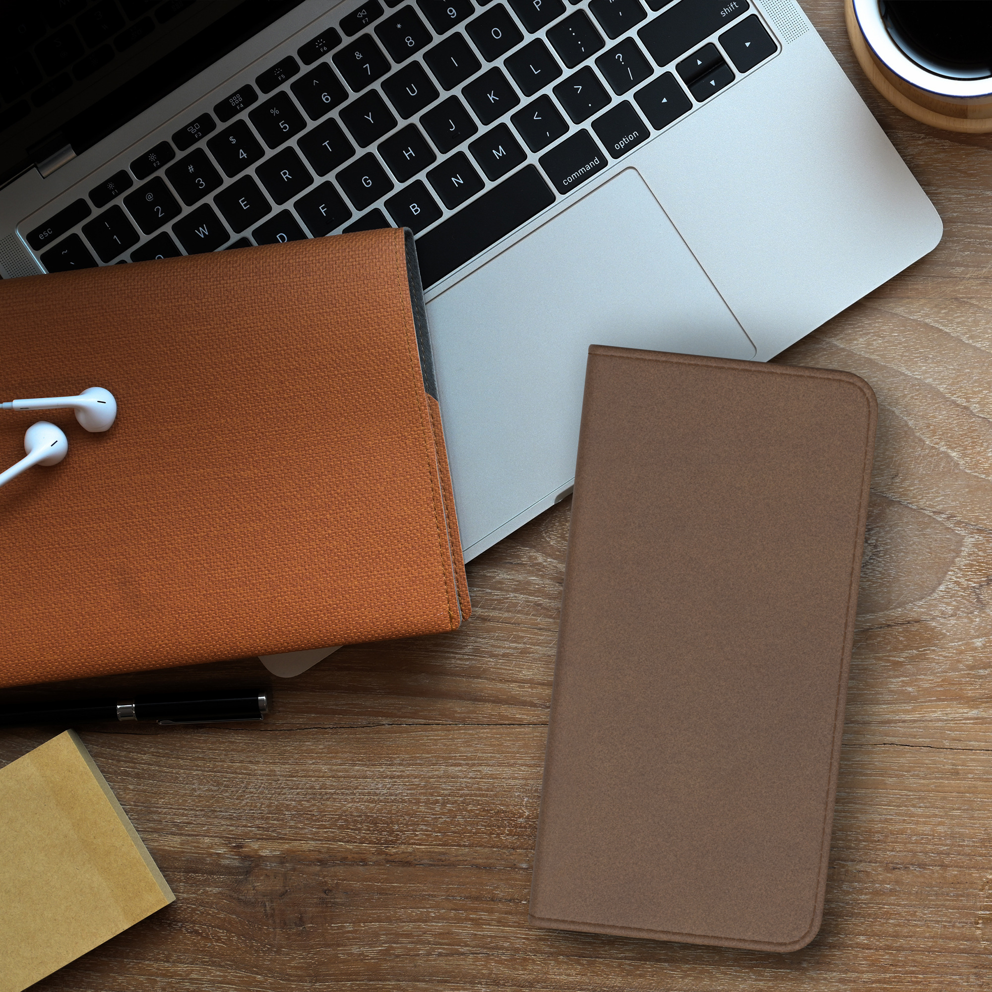 Ein Laptop, eine Handyhülle, ein Notizbuch und ein Stift liegen auf einem Holztisch. Es gibt eine Tasse Kaffee.