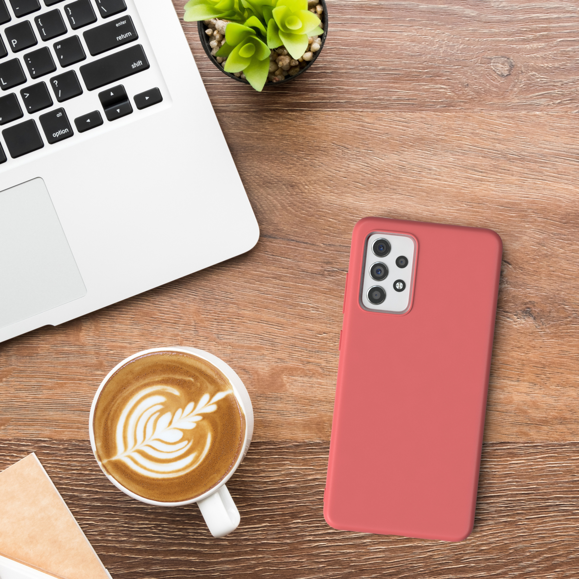 Smartphone in einer korallenfarbenen Hülle auf Holzoberfläche, mit Laptop, Kaffee und Pflanze im Hintergrund.