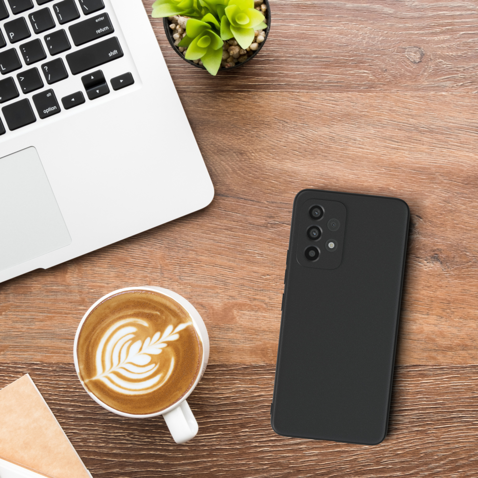 Ein Laptop, eine Tasse Kaffee, eine kleine Pflanze und ein schwarzes Telefon mit Hülle auf einem Holztisch.