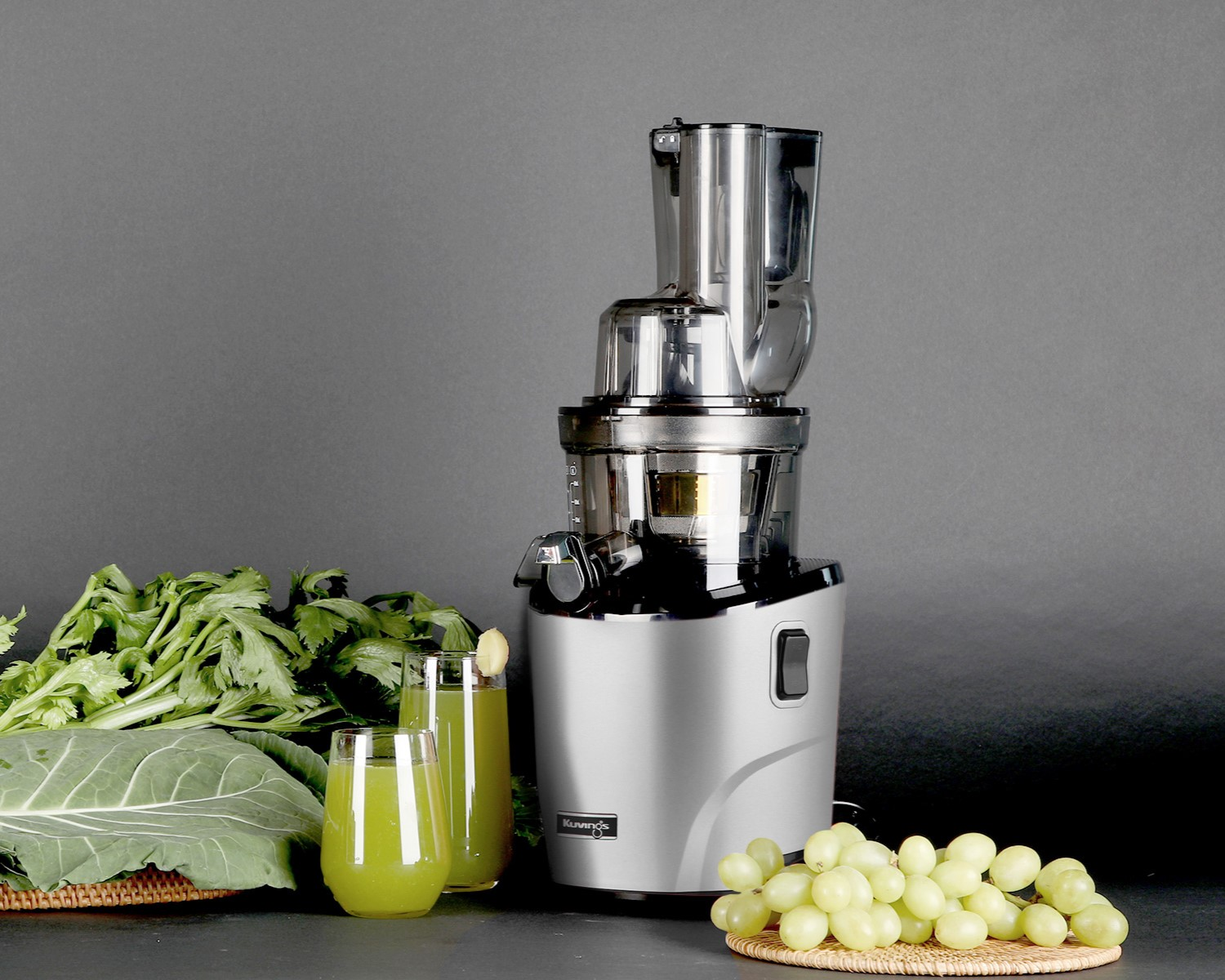Una licuadora, vasos con jugo y verduras, y un racimo de uvas en la mesa.