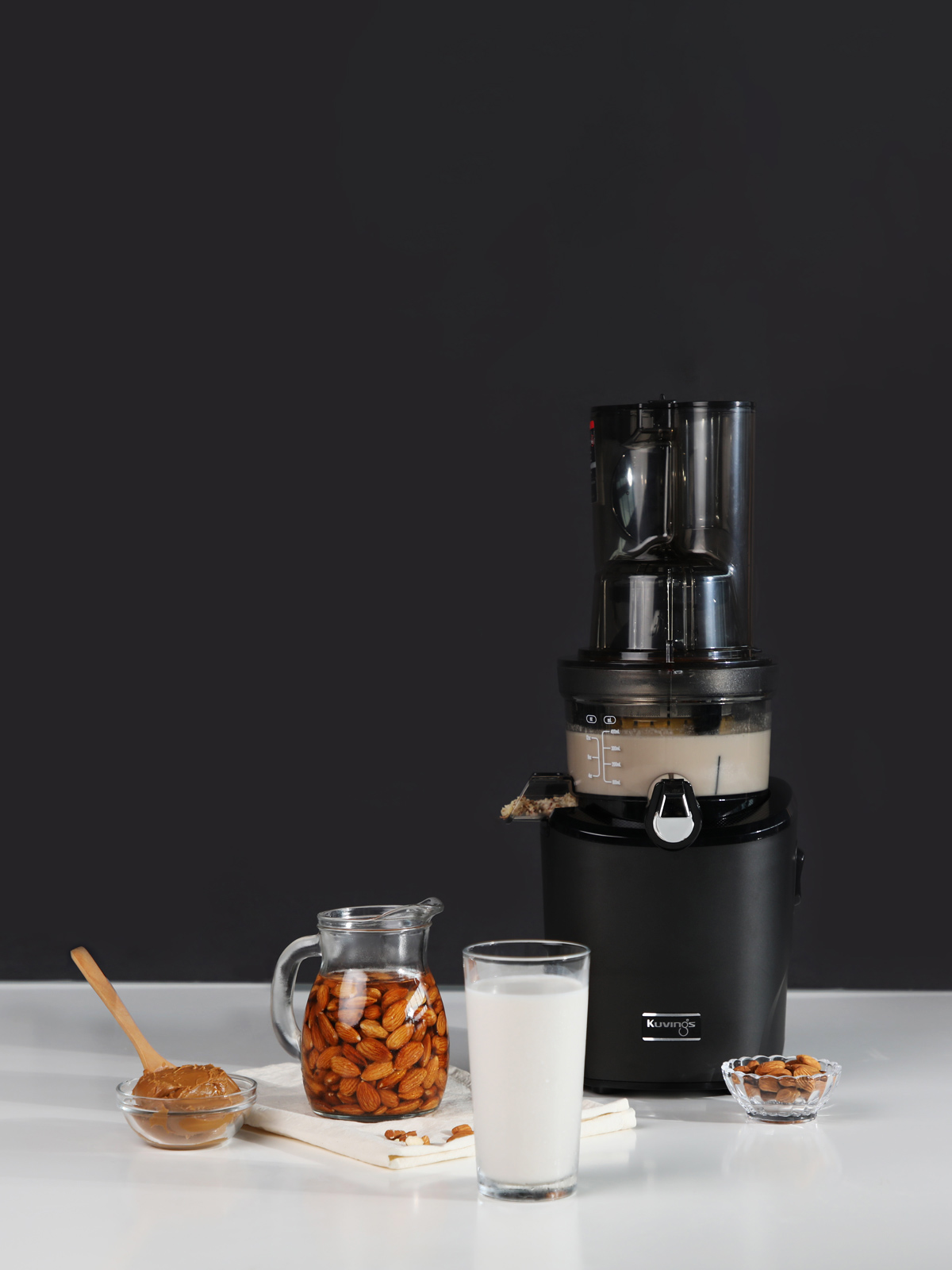 Exprimidor con almendras, leche de almendras y mantequilla de almendras. Fondo negro y superficie blanca.