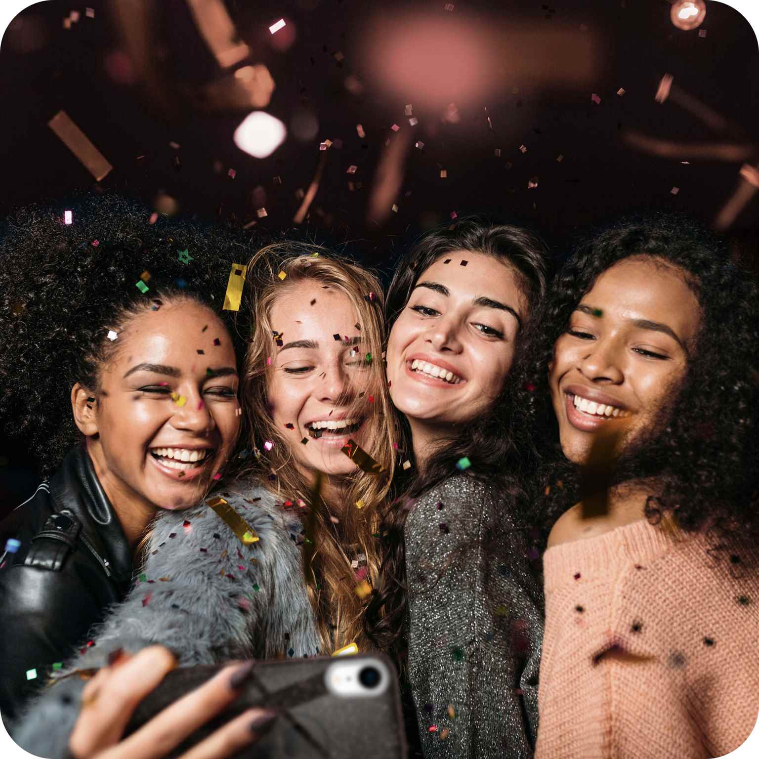 Vier lächelnde Frauen machen ein Selfie, Konfetti fällt in dunkler Umgebung um sie herum.