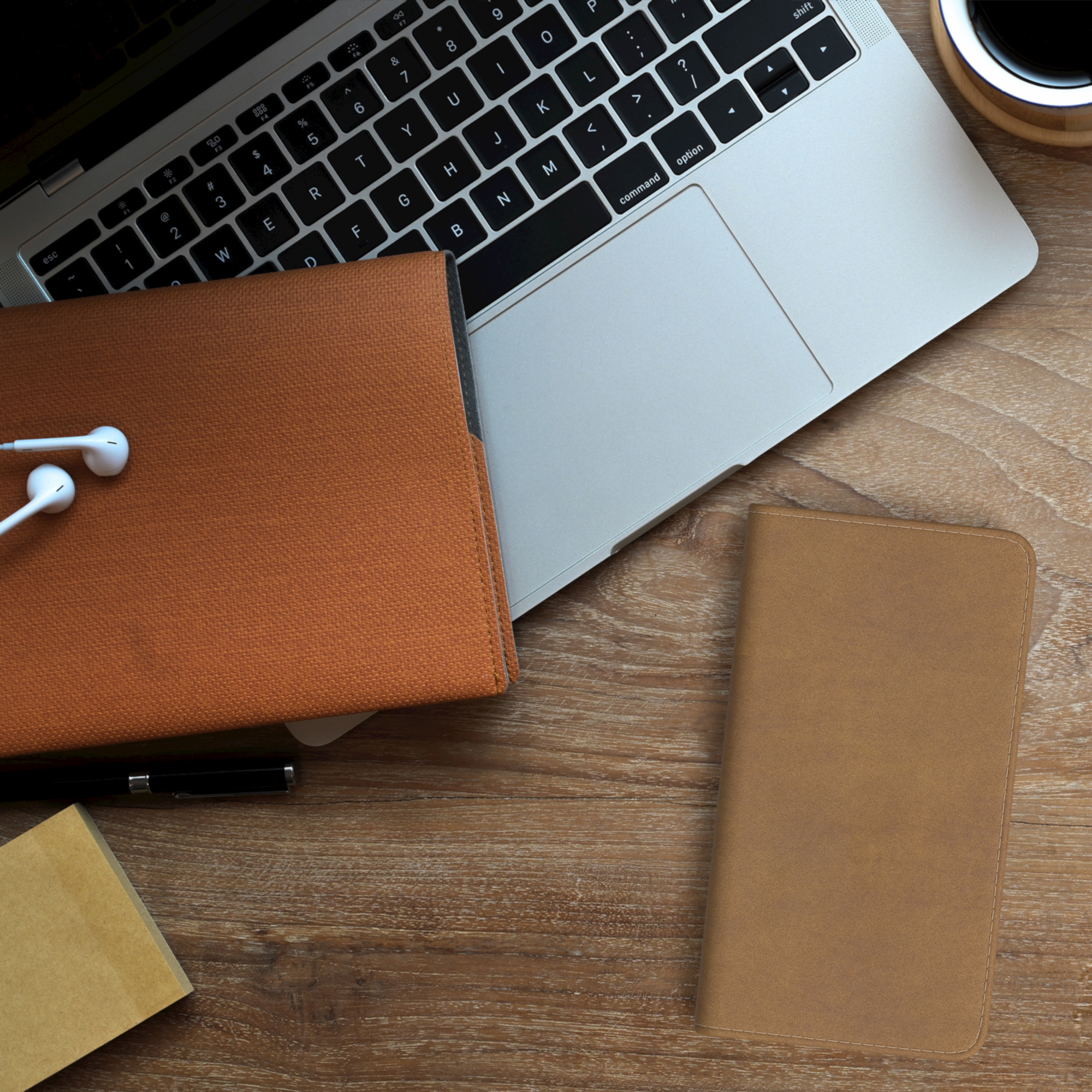Draufsicht auf einen Holzschreibtisch mit Laptop, Handyhülle, Kopfhörern, Notizblock, Stift und Kaffeetasse.