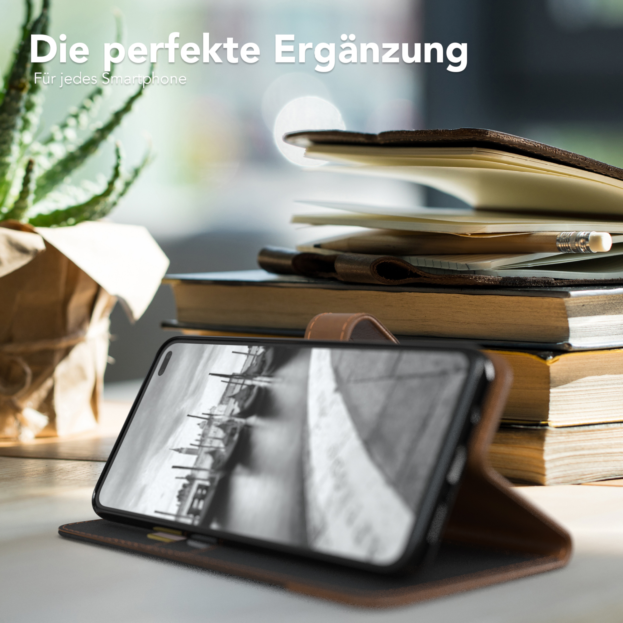 Telefon im Case, offen, auf einem Tisch, mit Büchern und einer Pflanze, der Bildschirm zeigt Stadt.