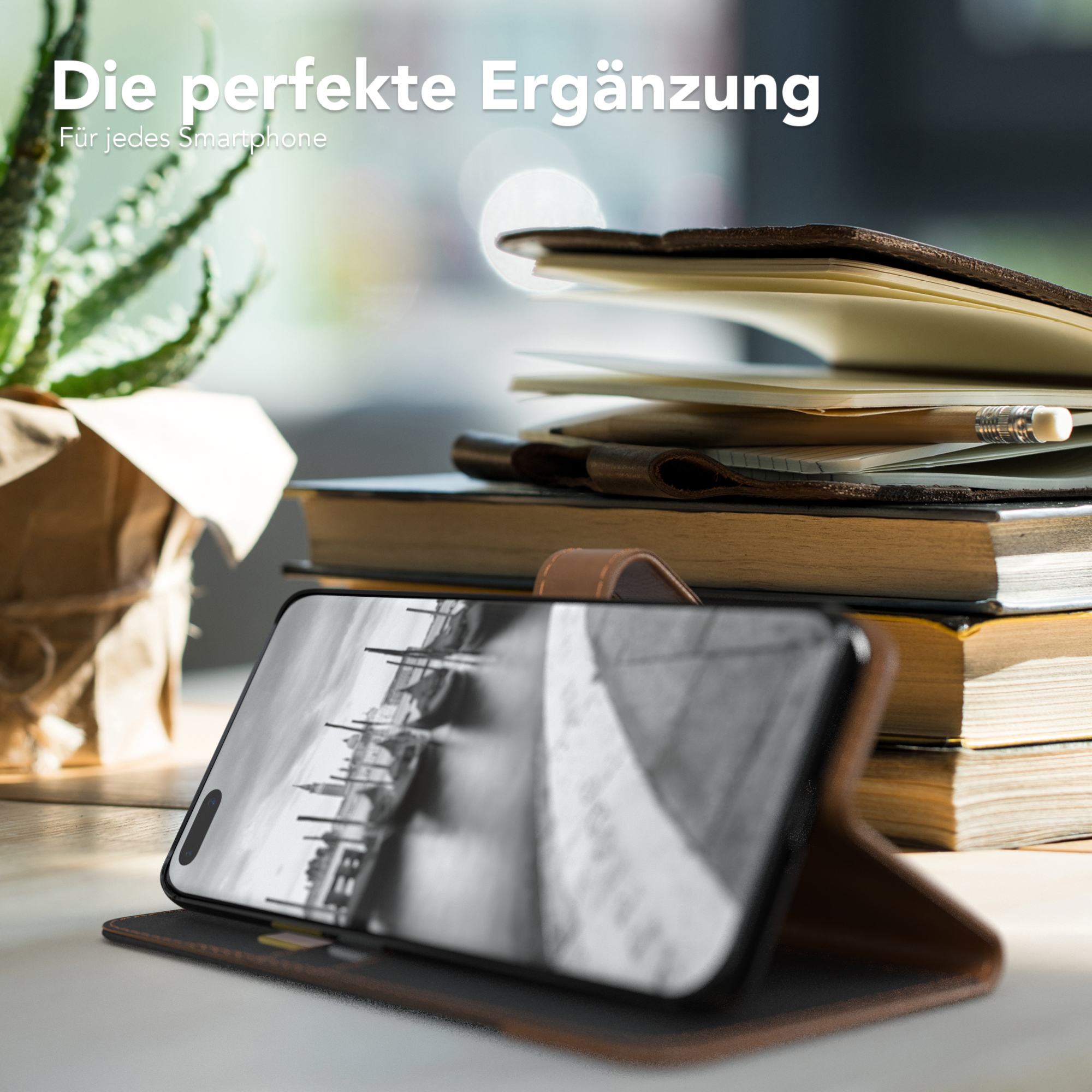 Telefon in braunem Etui auf einem Tisch, neben Büchern und einem Stift. Hintergrund unscharf.