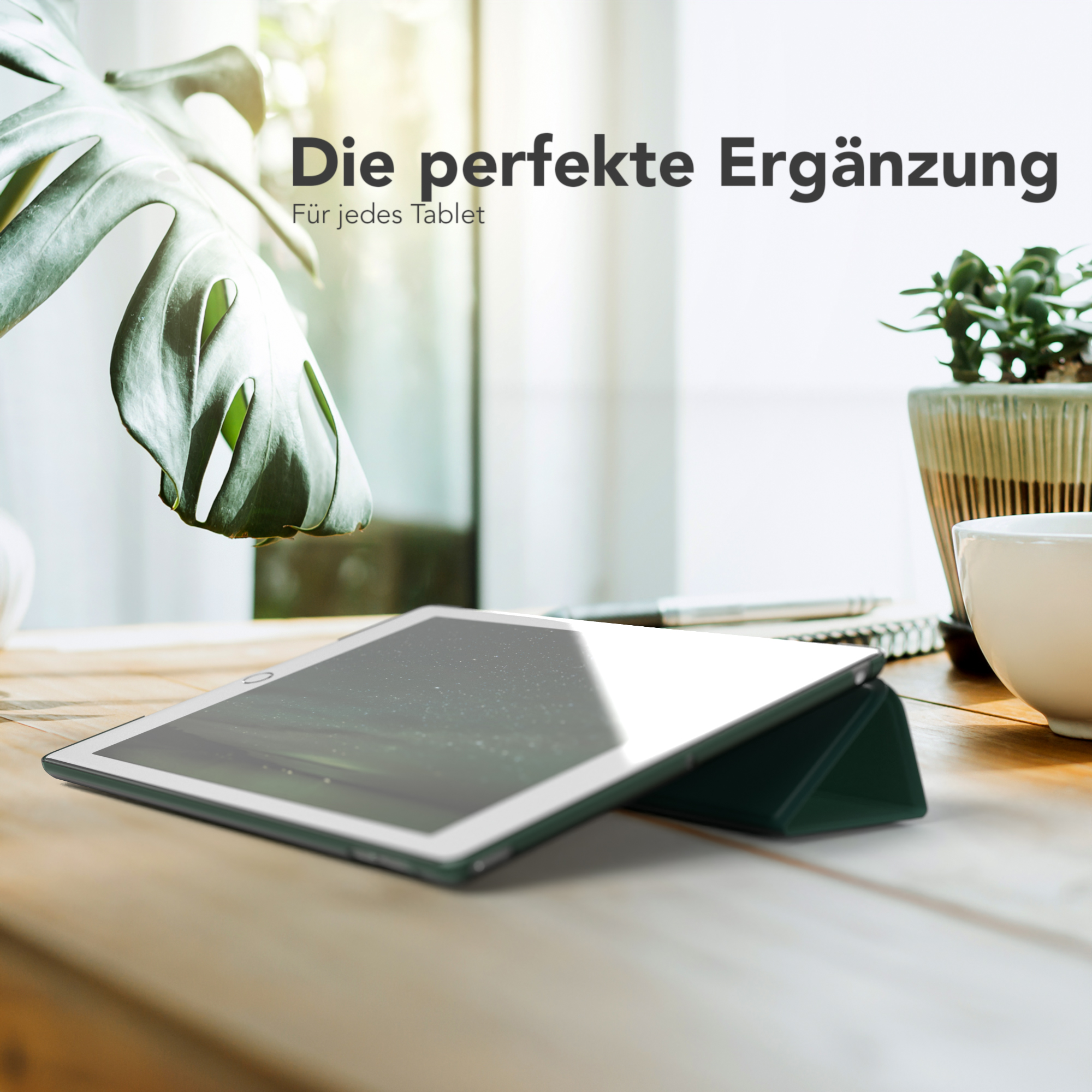 Tablet mit grüner Hülle liegt auf einem Holztisch. Eine Pflanze und eine Tasse sind im Hintergrund.