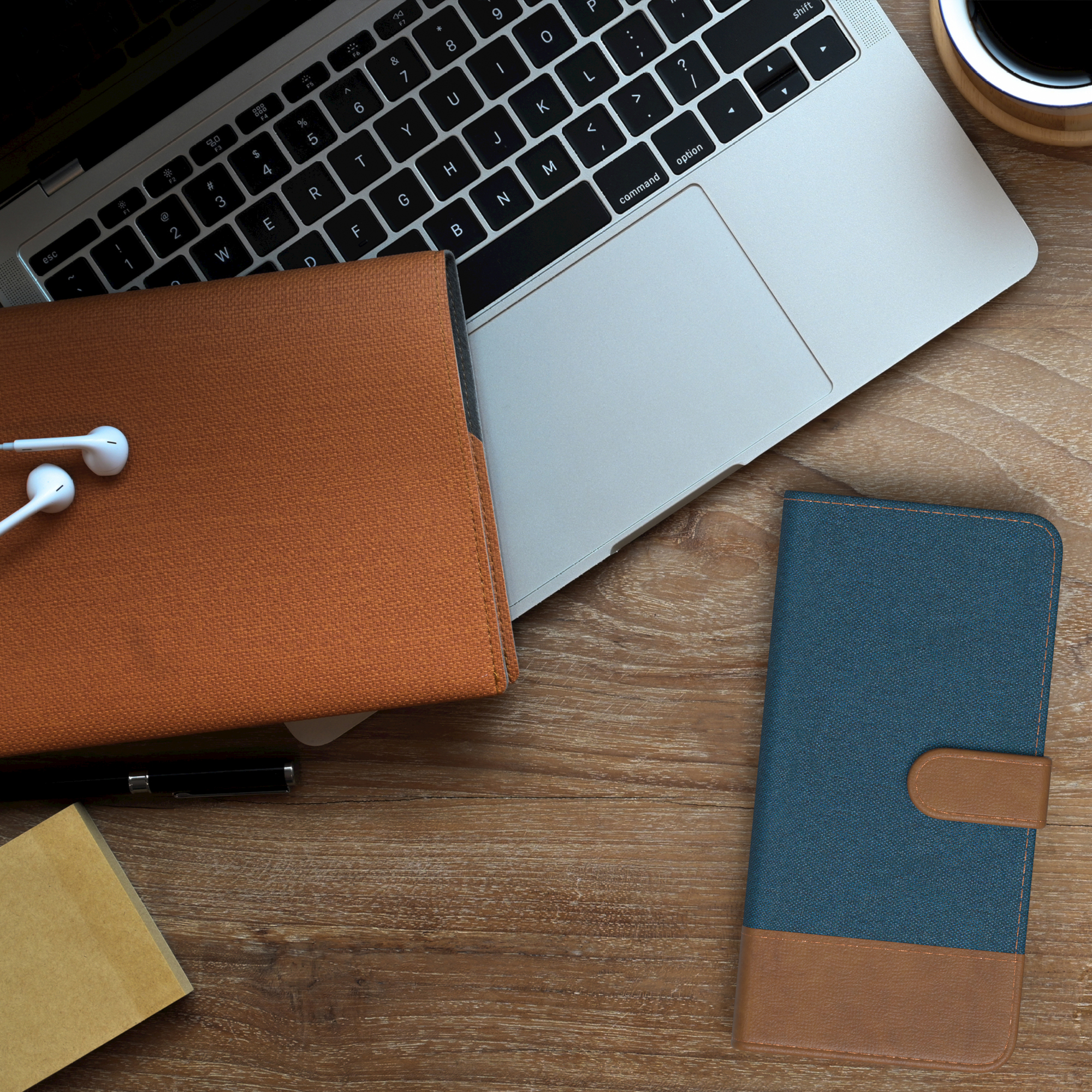 Laptop, Handyhülle, Notizblock, Kopfhörer, Stift und Kaffeetasse auf einem Holztisch.