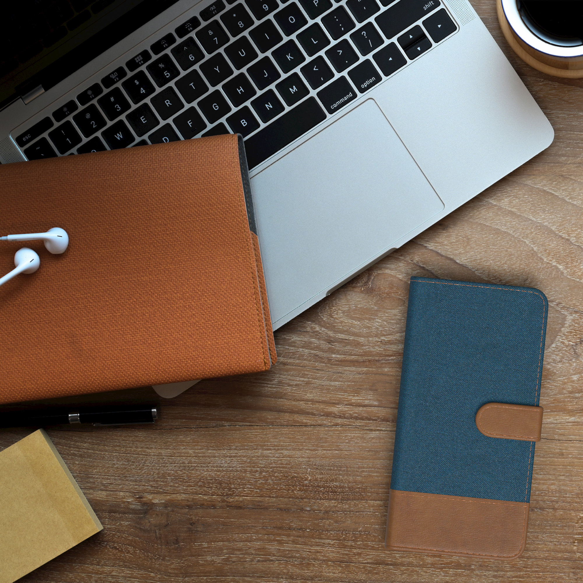 Laptop, Kopfhörer, Notizbuch und Handyhülle auf einem Holztisch. Kaffeetasse sichtbar. Arbeits- oder Lernumgebung.