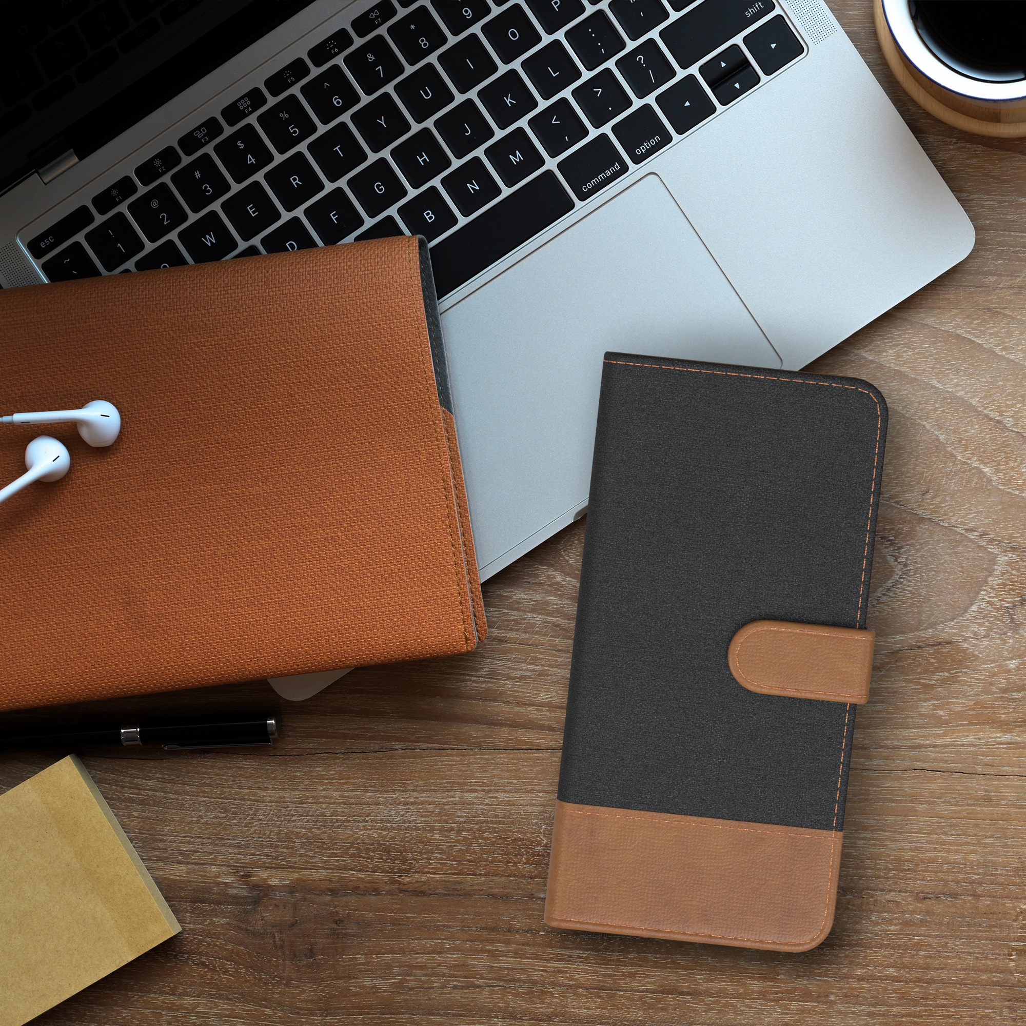 Ein Holztisch mit Laptop, brauner Hülle, schwarzer Handyhülle, Notizblock und Kaffeetasse.