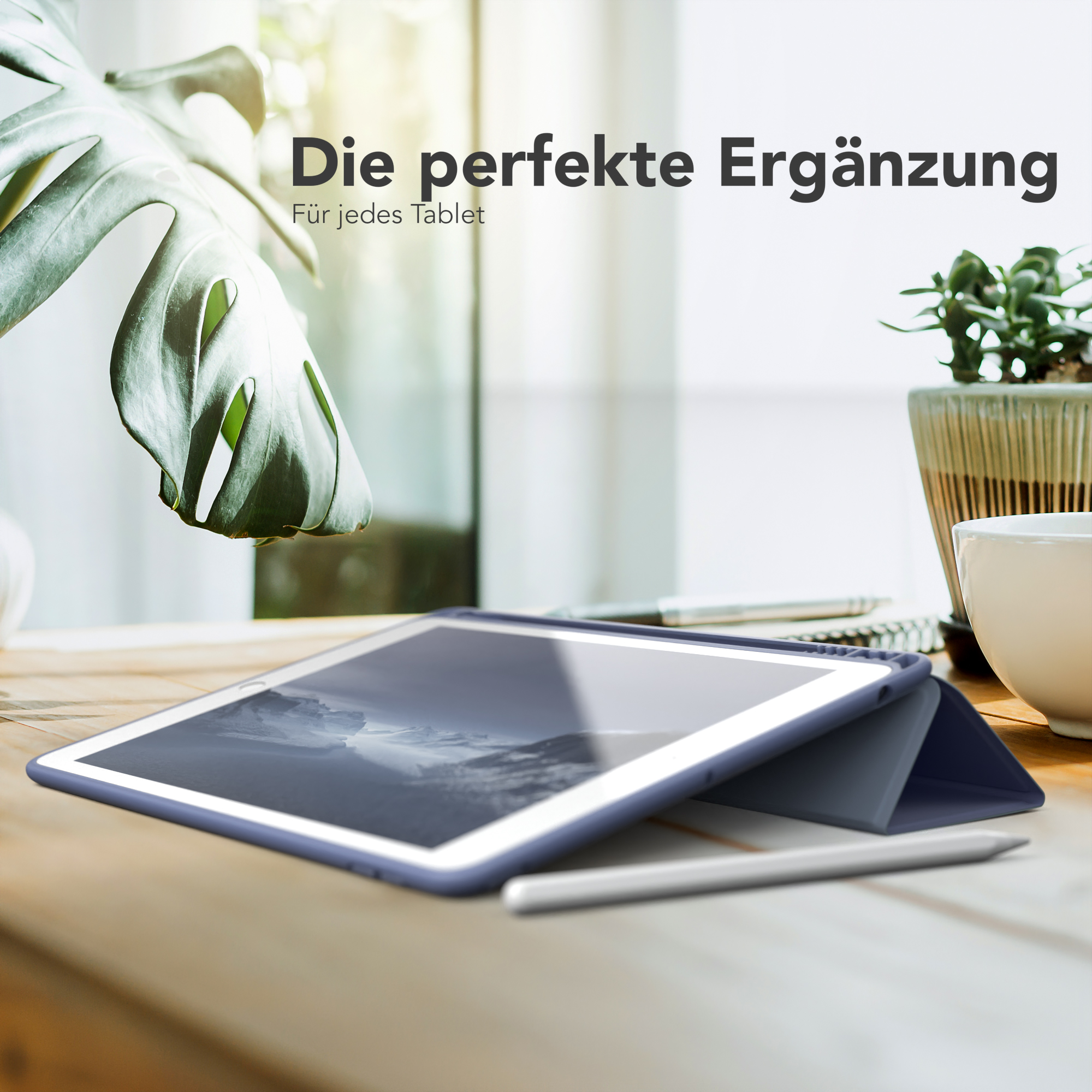Ein Tablet in einer Hülle und ein Stift liegen auf einem Tisch. Eine Pflanze und eine Tasse sind im Hintergrund.