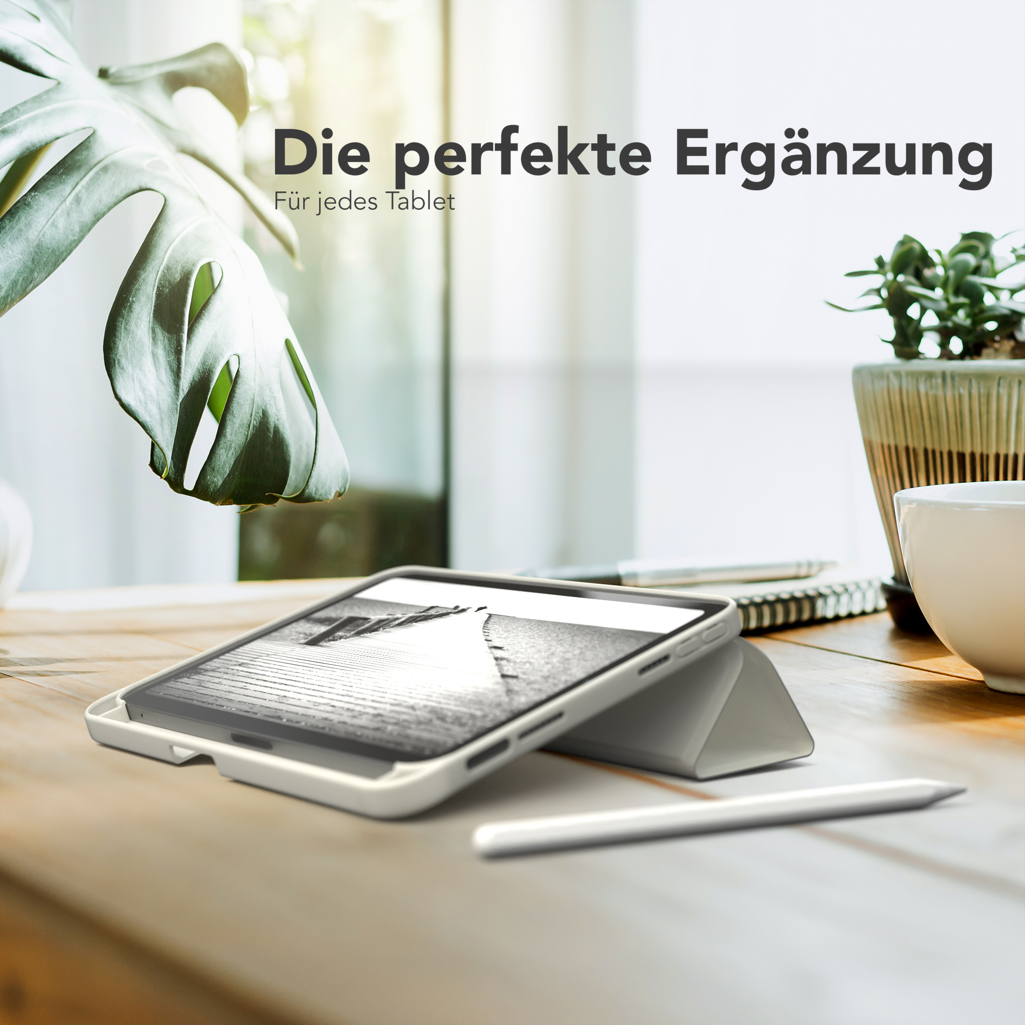 Tablet in weißem Gehäuse, mit Stift, auf Holztisch. Pflanze und Notizbuch im Hintergrund.
